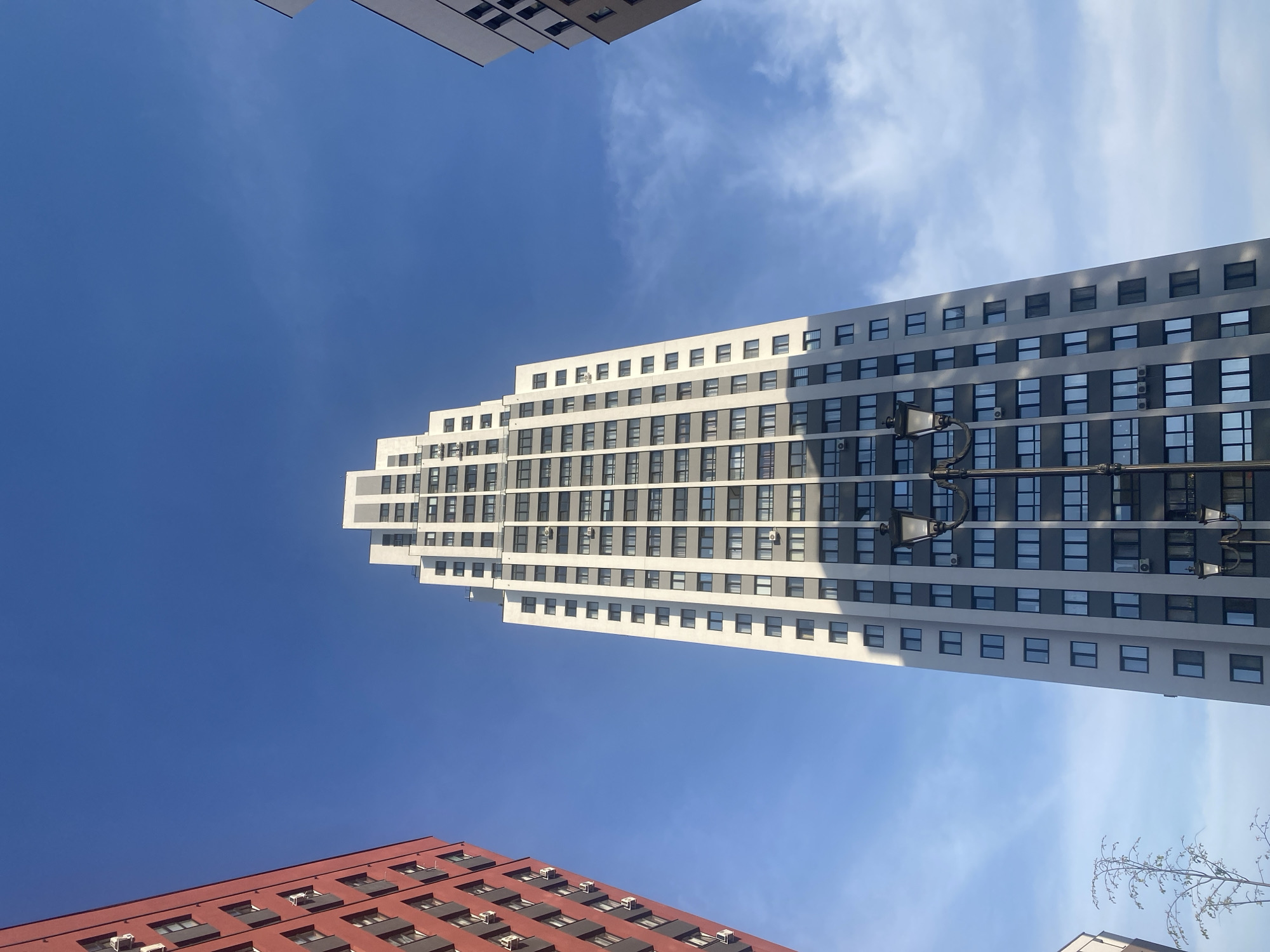 A low-angle, upward-facing shot features several buildings against a backdrop of a blue sky with scattered clouds.
The most prominent building, centrally located, is a tall, white skyscraper with numerous windows. The building tapers slightly towards the top.
To the lower left of the skyscraper is a red building with similarly designed windows. At the top left corner, a portion of a gray building is visible. A light pole is attached to the skyscraper. A bare tree branch is visible at the bottom right.
The sky transitions from a deep blue on the left to a lighter, cloudier shade on the right. The scene appears to be taken in Yekaterinburg, Russia, based on the architectural style of the buildings.