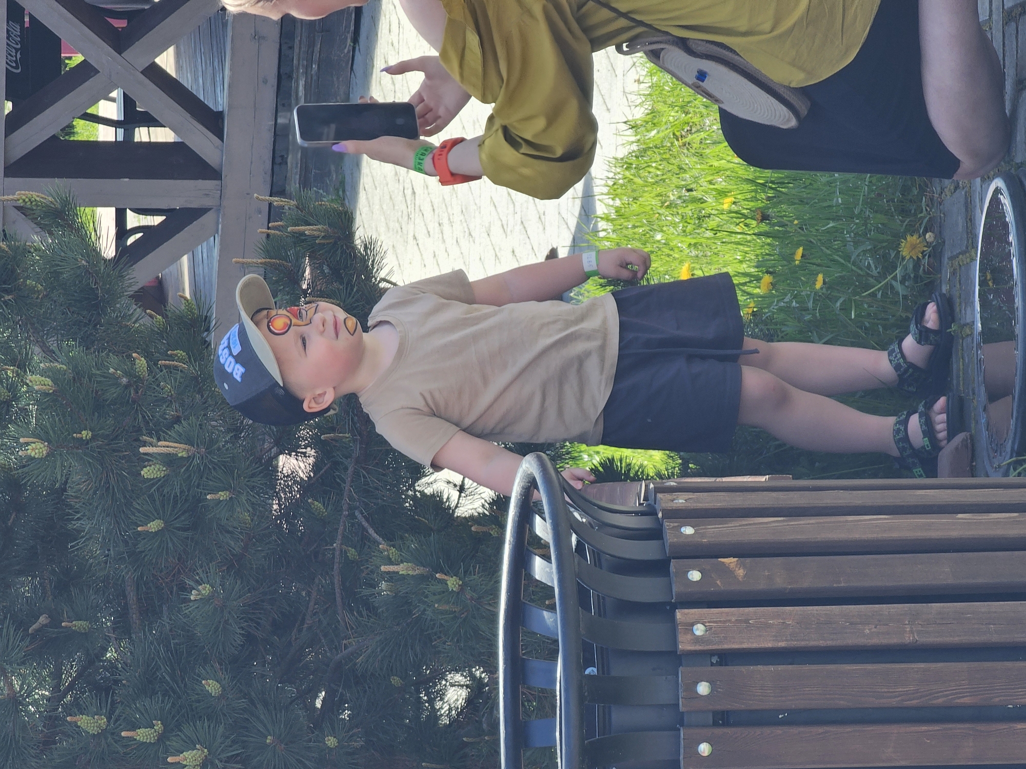 A boy with face paint is lying on a wooden bench next to a coniferous tree. The boy is wearing a tan shirt, blue shorts, and sandals. A woman is standing nearby, taking a picture of the boy with her phone. The location is inferred to be in Khimki, Russia.
