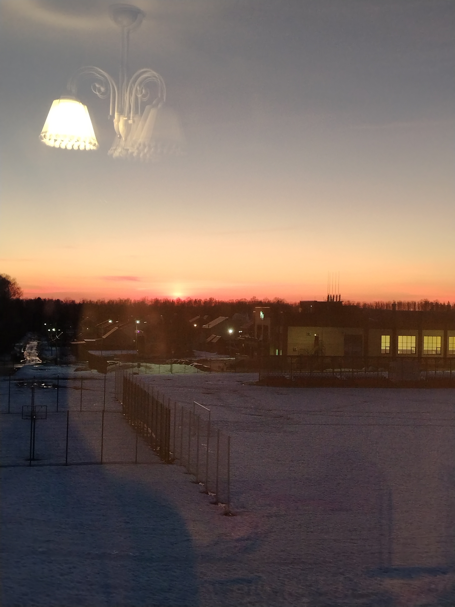 The image shows a snowy outdoor scene, likely taken through a window. The foreground consists of a snow-covered field or area with a fence running across it and several basketball hoops on the left. Beyond the fence, there is a row of trees and buildings with lights on, suggesting a town or urban area. In the background, the sky is a gradient of colors, ranging from a light grey at the top to a reddish-orange near the horizon, indicating a sunset or sunrise.
Superimposed on the scene in the upper portion of the image is a blurred chandelier, likely a reflection from the window through which the photo was taken. The overall environment suggests the location is Noviy, Russia.