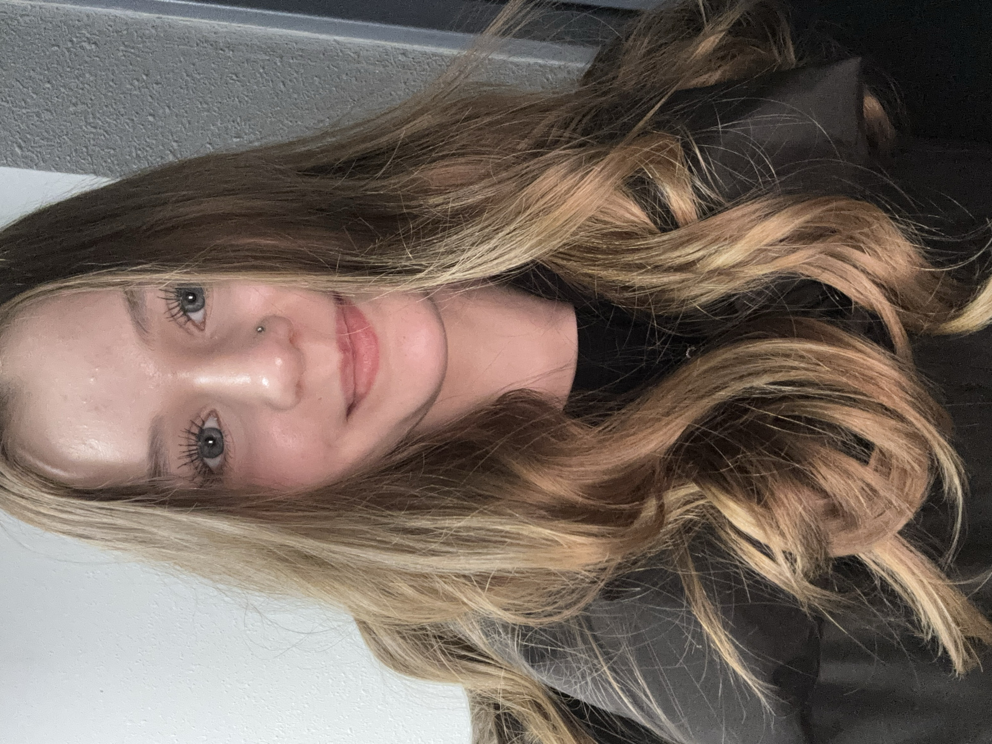 A close-up portrait of a woman with long, wavy, blonde and brown hair. She has light skin, blue eyes, and a small piercing in her nostril. She is wearing a dark-colored garment, possibly a jacket or shirt. The background consists of a plain white and grey wall. The image is oriented vertically.
There is no information available in the image to place the subject in Yekaterinburg, Russia.