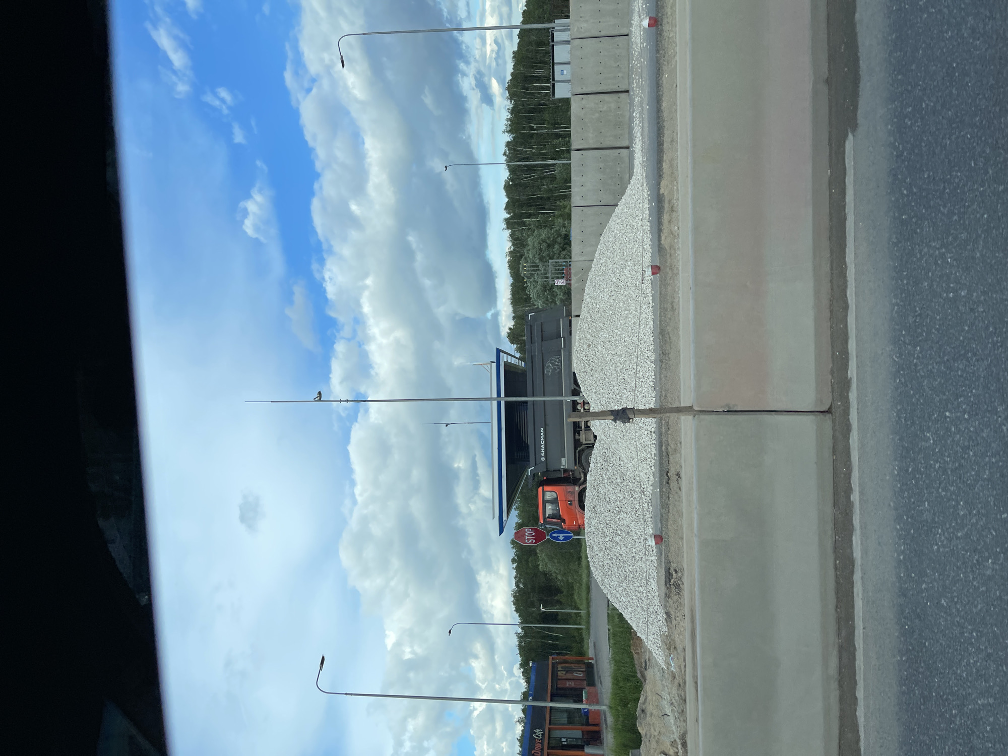 The image depicts a scene in Bykovka, Russia, showing a roadside view. A gray construction truck is visible. A pile of gravel is along a concrete barrier next to an asphalt road. The sky is blue with scattered white clouds. Utility poles are present. A Drive Cafe is also visible in the lower part of the image. A stop sign is next to the construction truck. A forested area is in the background.