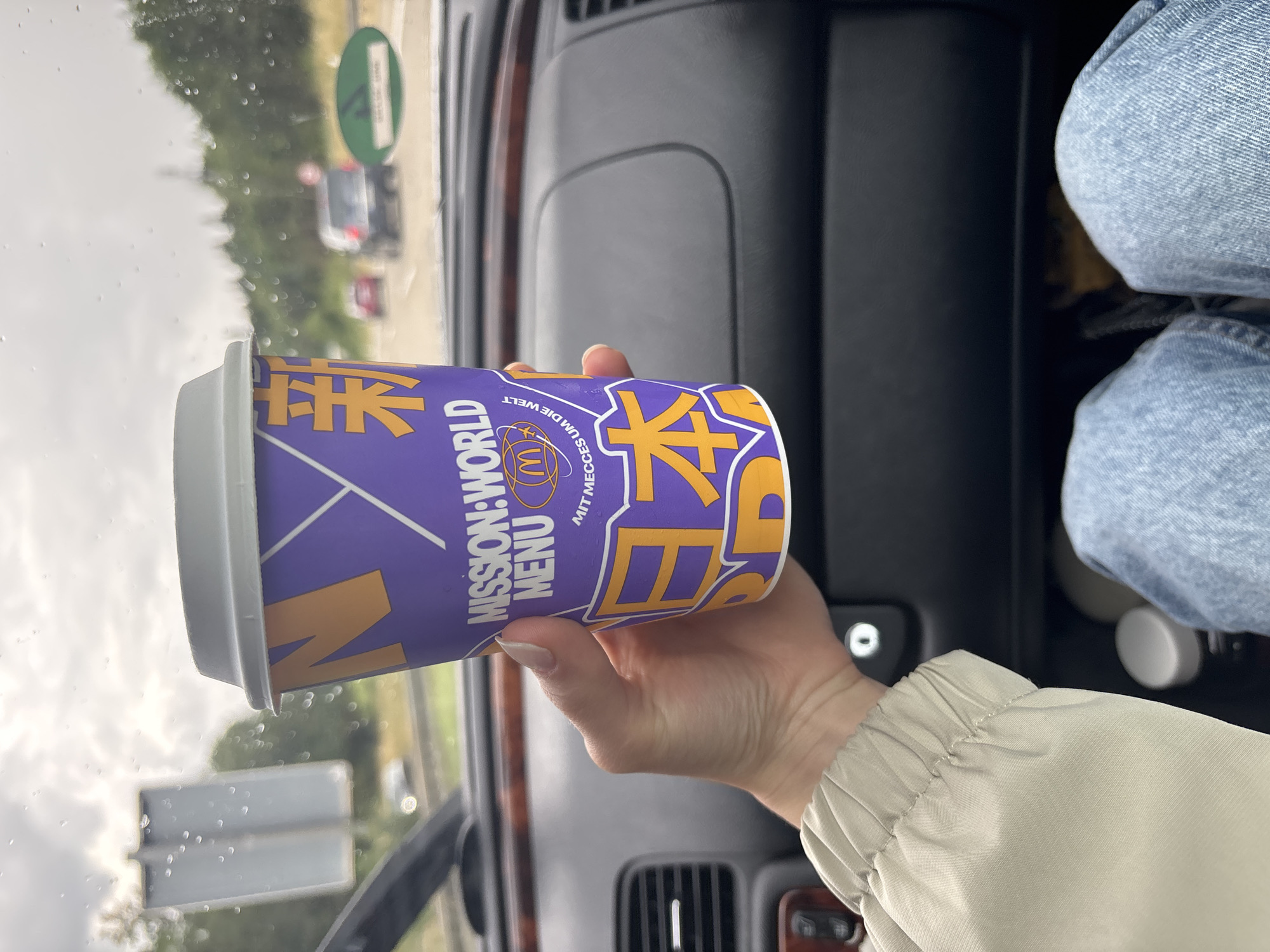 A person holds a purple McDonald's "Mission: World Menu" cup in their hand. The cup features orange lettering and graphics, including what appear to be Japanese characters. The background shows the interior of a car, with a dashboard and the windshield visible. Outside the car, a road with some cars and trees can be seen, suggesting the car is in motion. The sky is overcast. The image was taken in Aalen, Germany.