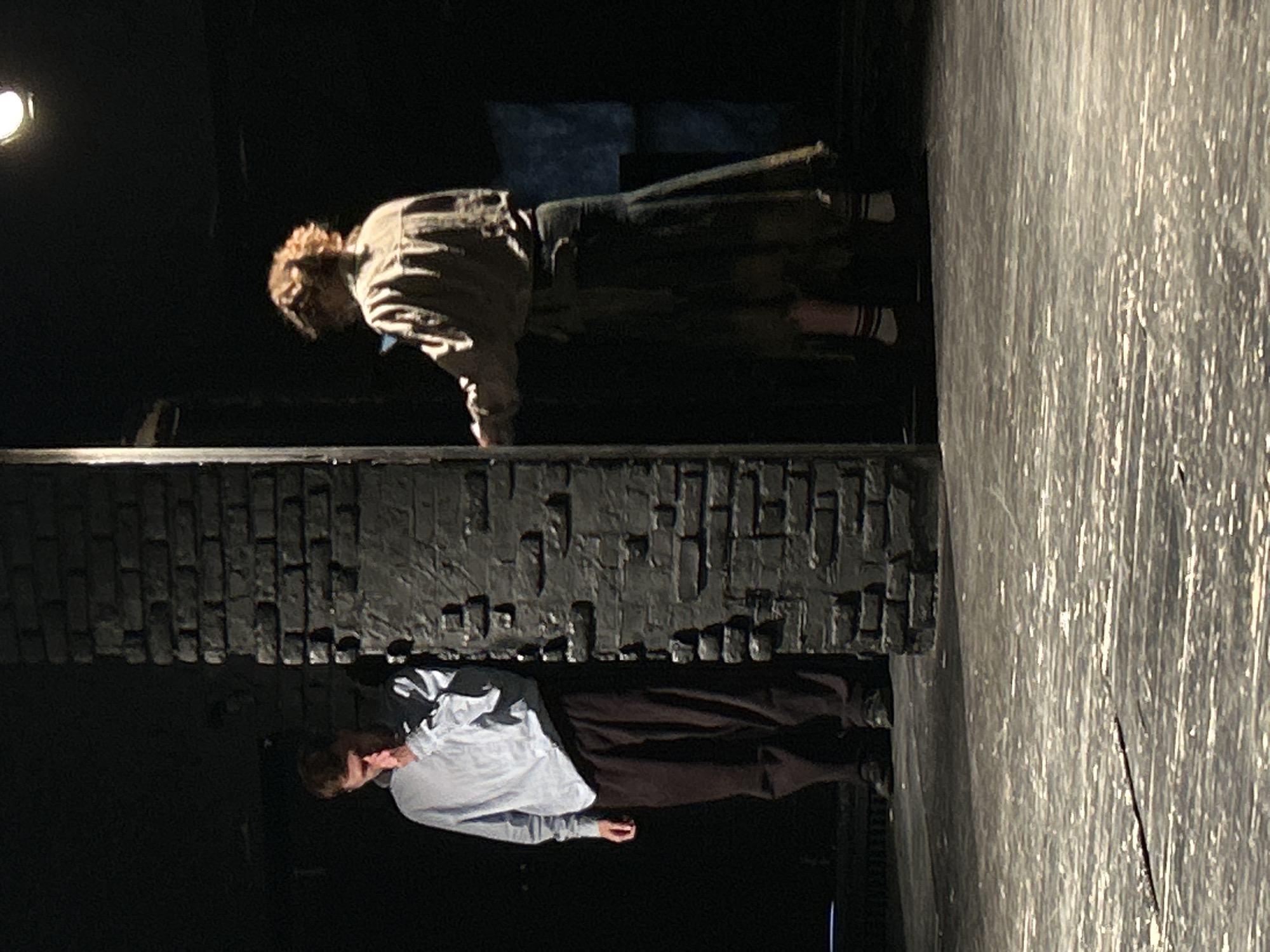 The image captures a theatrical performance in a darkened setting. A brick-like structure divides the scene horizontally. Above the structure, a figure with blonde hair, wearing a grey jacket and dark kilt or skirt with striped socks, leans forward. Below the structure, another figure in a light blue shirt and dark trousers appears to be positioned prone. The environment consists of a dark background and a stage floor with what appears to be light patterns. The exact location in the city of Unknown, Unknown cannot be determined without additional information.