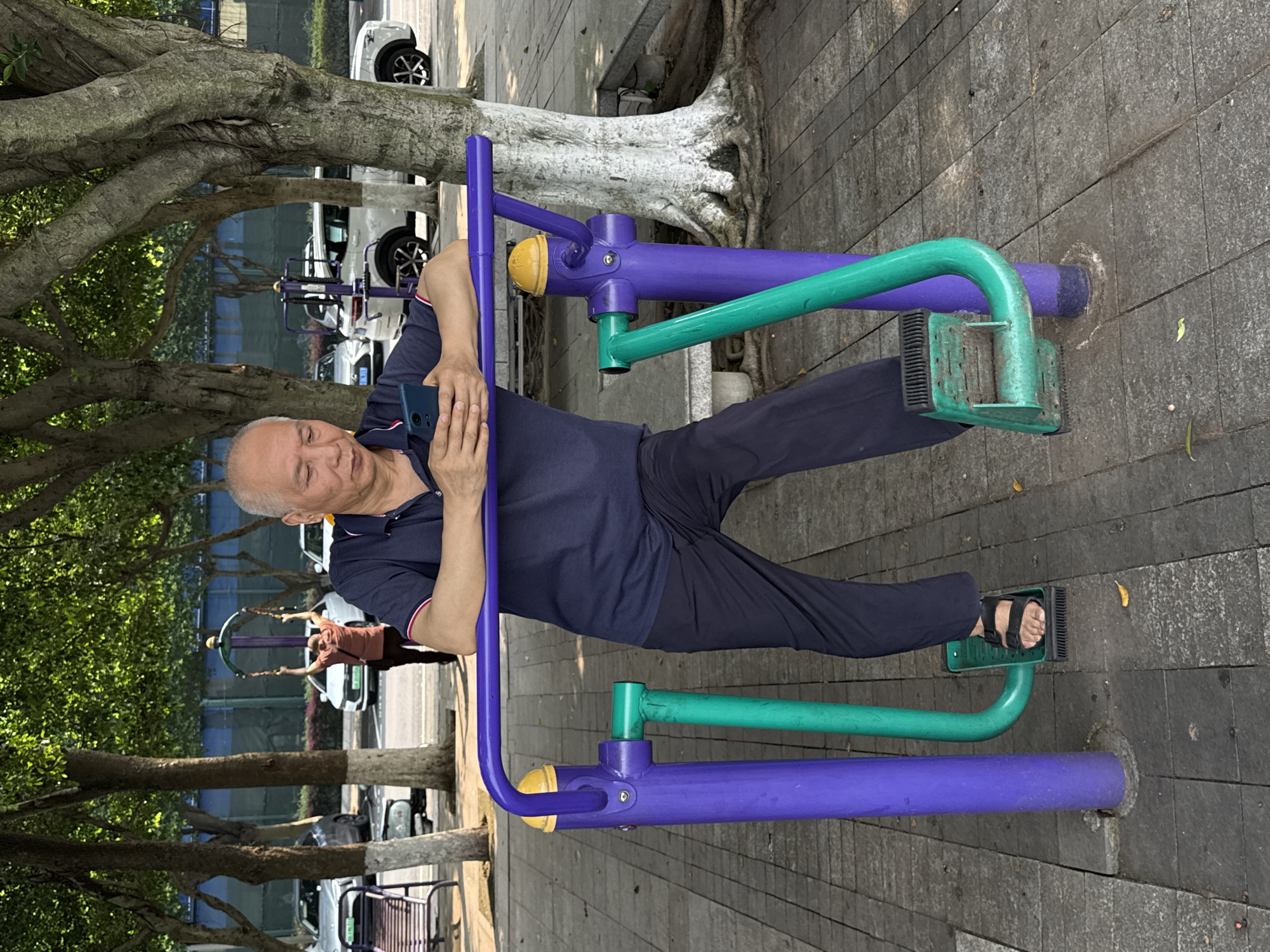 An elderly man is standing on outdoor exercise equipment in a public park in Jiangjin, China. He is positioned facing slightly left of the camera, wearing a dark blue polo shirt with red and white trim on the sleeves, and dark blue trousers. On his feet are black sandals. His forearms rest on a purple horizontal bar, and his hands are occupied with a dark blue smartphone, which he is looking down at. The exercise equipment features two vertical purple posts, one on each side, connected by the horizontal bar. Yellow caps top the vertical posts. Green curved metal bars extend downwards from the purple posts, ending in green foot pedals with black textured surfaces, one of which the man's right foot occupies. The ground is paved with rectangular grey tiles.
In the mid-ground, behind the man and to his left, another individual is seen from behind, engaged with a different piece of outdoor exercise equipment, raising their arms above their head. This equipment is also purple and green. Numerous deciduous trees with light grey bark and lush green foliage are scattered throughout the scene, casting dappled sunlight. Beyond the trees, a paved road is visible, with several white and light-colored cars parked or driving. A blue structure or wall and green fencing are discernible further in the background.