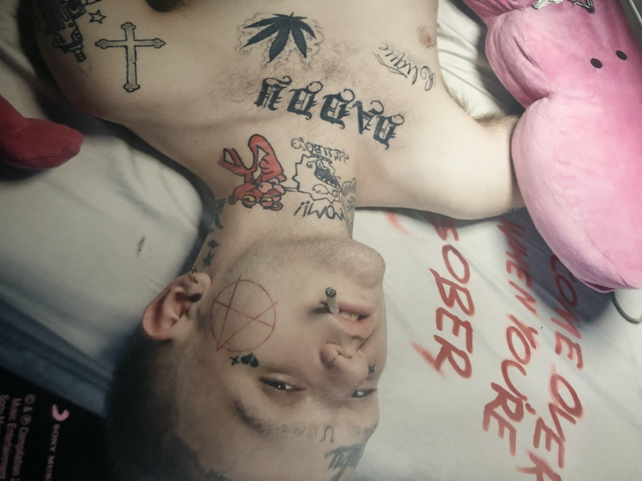 An overhead photograph captures a male subject, Gustav Åhr (Lil Peep), lying supine on a white surface, likely a bed, with his head slightly angled to his left. His eyes are open, and a lit cigarette is held between his lips through a labret piercing. The subject's face, neck, and upper torso are extensively tattooed. Facial tattoos include a red outlined anarchist 'A' symbol on his right cheek, a broken heart and 'X' under his left eye, and "crybaby" above his right eyebrow. A nose ring is also present. Prominent neck tattoos feature a red Pink Panther character on the right side and a cartoon character resembling Betty Boop on the left. His upper chest displays a large, gothic-style "DADDY" tattoo centrally, a cannabis leaf to its right, and a prominent cross to his upper right. Other smaller tattoos like "oblique" and "GOTHBΟΥ" are visible. The white surface beneath him is marked with large, red, stylized letters forming the phrase "COME OVER WHEN YOU'RE SOBER," oriented to be read from bottom to top. A pink plush object, possibly a heart-shaped pillow, is partially visible at the bottom right. In the upper left background, a portion of a black object with white text is visible, including "SONY Music Entertainment" and a copyright date of "2017." The image contains no visual identifiers to establish a specific location within the city of Paris, France.