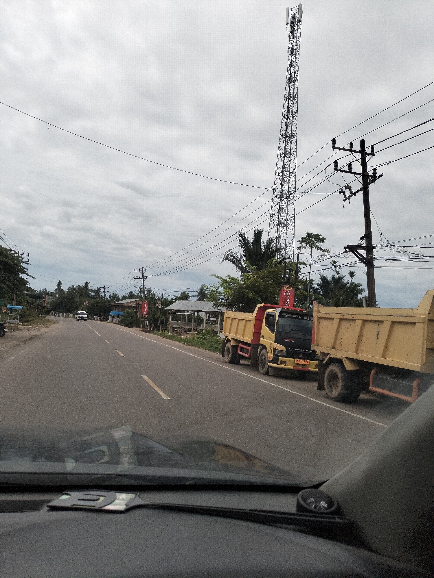 A frontal, slightly angled view from inside a vehicle shows a two-lane asphalt road in Matangkruet, Indonesia. The sky is overcast with grey clouds. On the right side of the road, there are two yellow dump trucks parked side-by-side. The truck in the foreground is closer to the viewer, showing its rear section. The truck behind it is mostly visible, including its front cabin which has "MITSUBISHI FUSO" written on it. The license plate reads "IN 4512 HG".
Beyond the trucks, a tall, lattice-style cell tower stands prominently against the sky. Next to it, a wooden utility pole with multiple wires crisscrossing the scene supports electrical lines. Further down the road, on both sides, are various structures including what appear to be small buildings or stalls with corrugated metal roofs, surrounded by lush green trees and palm trees. A white van is visible in the distance on the left side of the road. The road itself has faded yellow dashed lines marking the lanes. The interior of the vehicle is partially visible in the foreground, showing part of the dashboard and windshield.