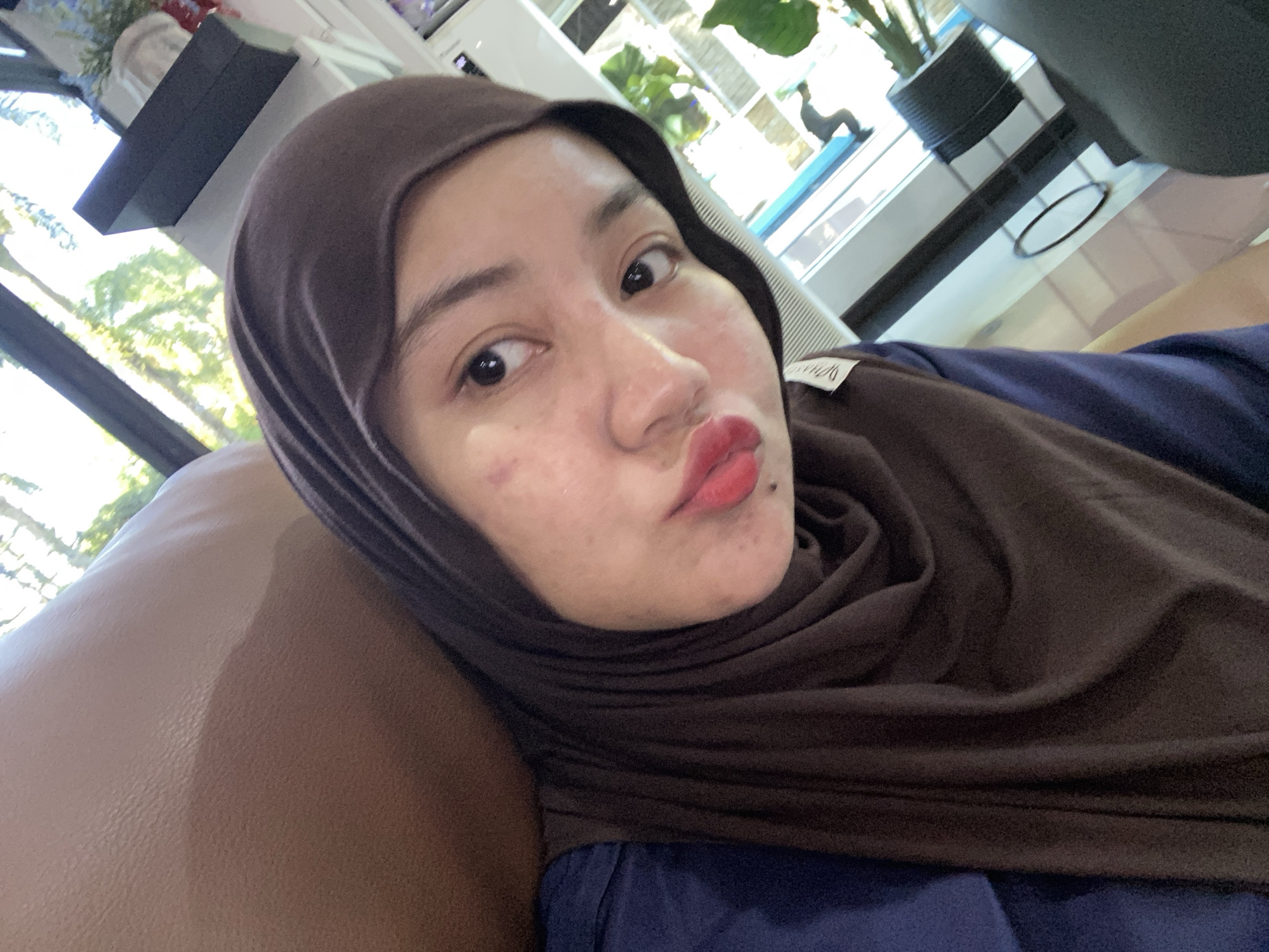 A young woman wearing a brown hijab and a blue top takes a selfie in a brightly lit indoor setting, possibly a cafe or lounge, in Banjarbaru, Indonesia. Sunlight streams in from large windows, illuminating trees and foliage outside, suggesting a daytime scene. The woman's face, with visible blemishes and bright red lipstick, is the primary focus. She is seated on a brown, textured surface, possibly a sofa or chair. The background shows hints of indoor furnishings and reflective surfaces, indicating a public or commercial space.