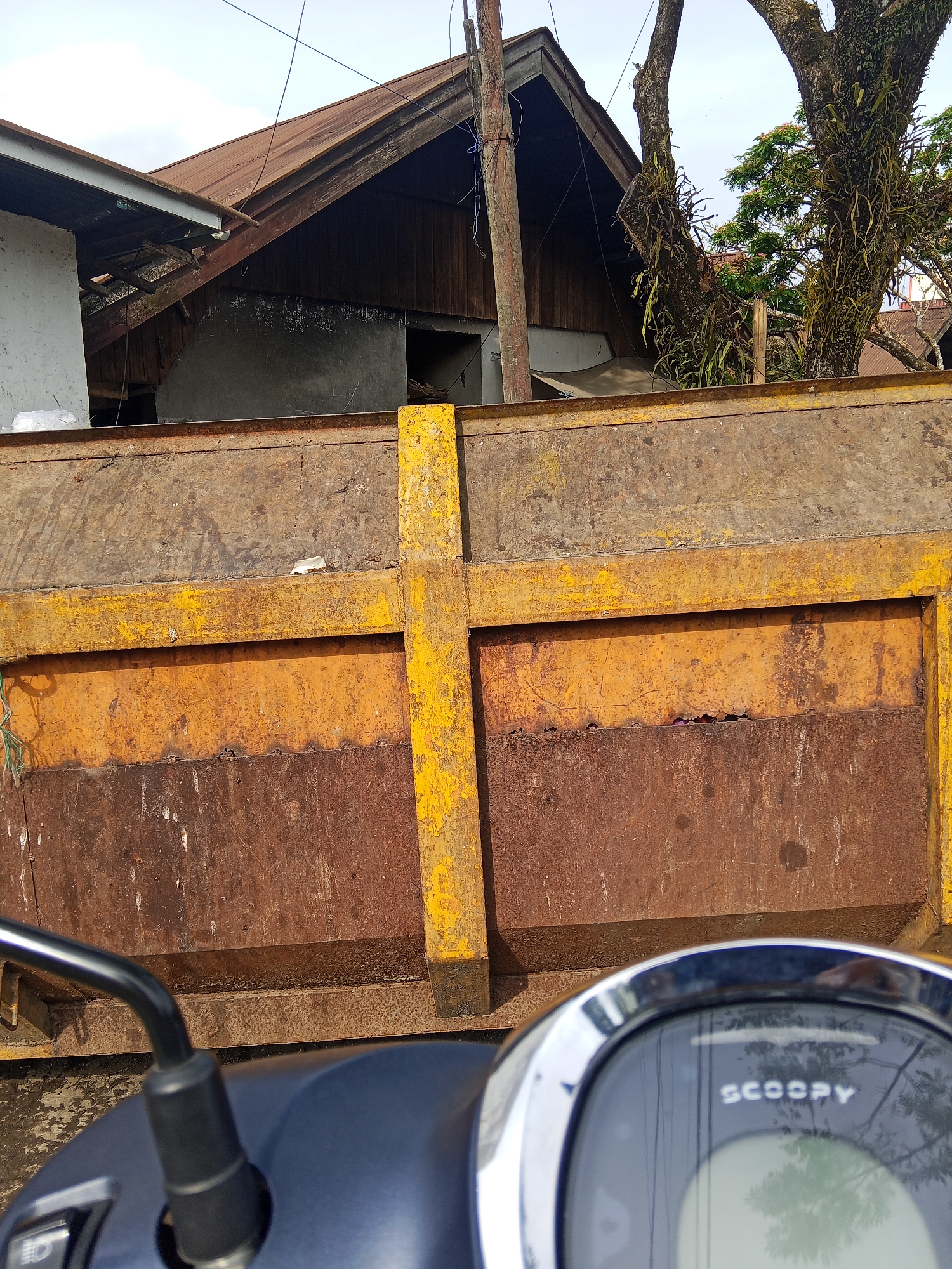 In Makale, Indonesia, the foreground of the image is dominated by the blue dashboard and a chrome-rimmed display of a scooter. Partially visible to the left is a black handlebar with a black mirror attached. Behind the scooter, a large, rusty metal structure, likely a truck bed, takes up the majority of the frame. This structure is a dull brown color with streaks of orange and yellow paint, indicating significant wear and tear. A prominent yellow vertical beam is bolted to the front of this structure, crossing a horizontal yellow beam. Beyond the metal structure, the background shows a dilapidated building with a weathered brown wooden roof and grey concrete walls. A large, old tree with moss and vines growing on its branches is visible to the right of the building, adding to the scene's rustic and somewhat neglected appearance. The sky above is a pale blue, suggesting it is daytime.