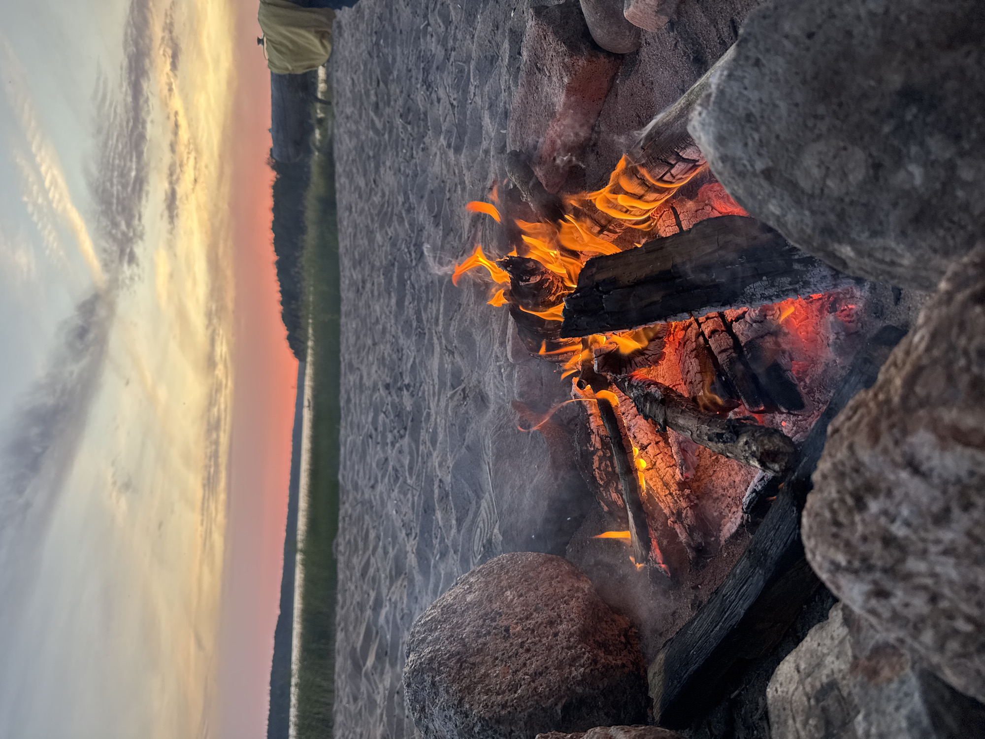 The image depicts a campfire on a sandy beach at dusk. The fire is burning brightly with visible flames and glowing embers, surrounded by rocks. In the background, a body of water is visible, with a tree line along the horizon. The sky displays a gradient of colors, from orange and pink near the horizon to pale blue and gray higher up, with scattered clouds. The sand shows footprints. A person in a yellow garment is partially visible in the far right background, near a log. The location is identified as Красная Долина, Russia.