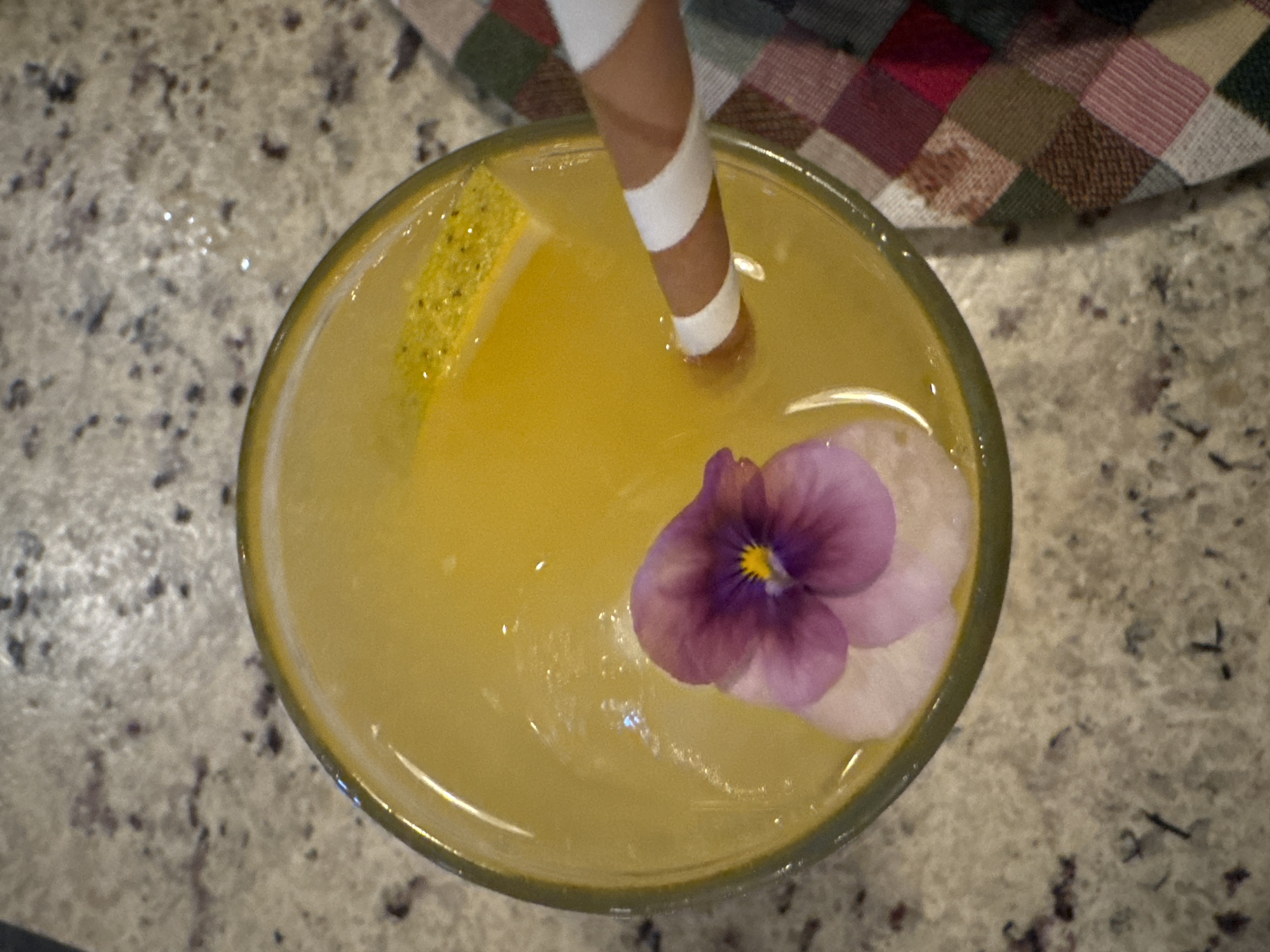 A high-angle shot depicts a yellow beverage in a glass, garnished with a purple flower and a lemon wedge. A striped straw is partially submerged in the drink. The glass sits on a speckled surface, and a checkered cloth appears in the background. No people are visible. The location is not discernible from the image.