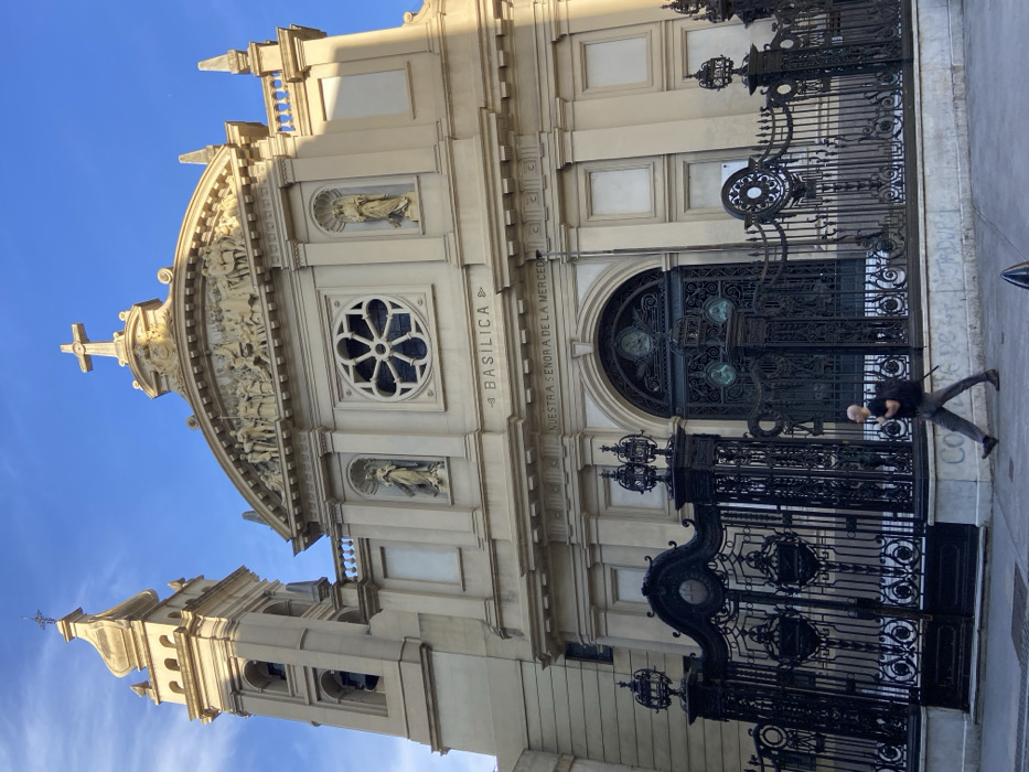 A rotated exterior shot of the Basilica Nuestra Señora de la Merced. Its ornate façade has classical features, including arched windows, statues, and a decorative rose window. A towering, black iron gate with intricate designs stands in front. A person walks past, casting a long shadow. The sky is partly visible, a clear blue.