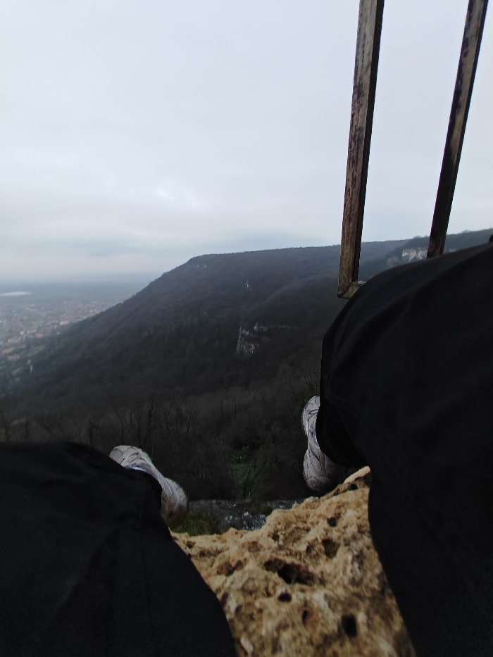 The image displays a first-person, downward-angled view from a high elevation, specifically from a rocky cliff edge overlooking the city of Shumen, Bulgaria. In the immediate foreground, a textured, light-colored rocky surface with visible pores and cracks occupies the bottom quarter of the frame. A person's lower legs are visible, clad in dark trousers and light-colored athletic shoes, with their feet suspended outwards over the cliff edge. Two weathered, dark vertical posts, likely part of a protective railing or observation structure, are present on the right side of the frame, extending upwards. The midground reveals a dense, dark forested mountain slope descending sharply, with numerous bare or dark-foliaged trees and some exposed rocky sections. In the distant background, the urban area of Shumen, Bulgaria, is partially visible through atmospheric haze, situated in a valley against further rolling terrain. The sky is uniformly overcast and grey, indicating a cloudy weather condition.