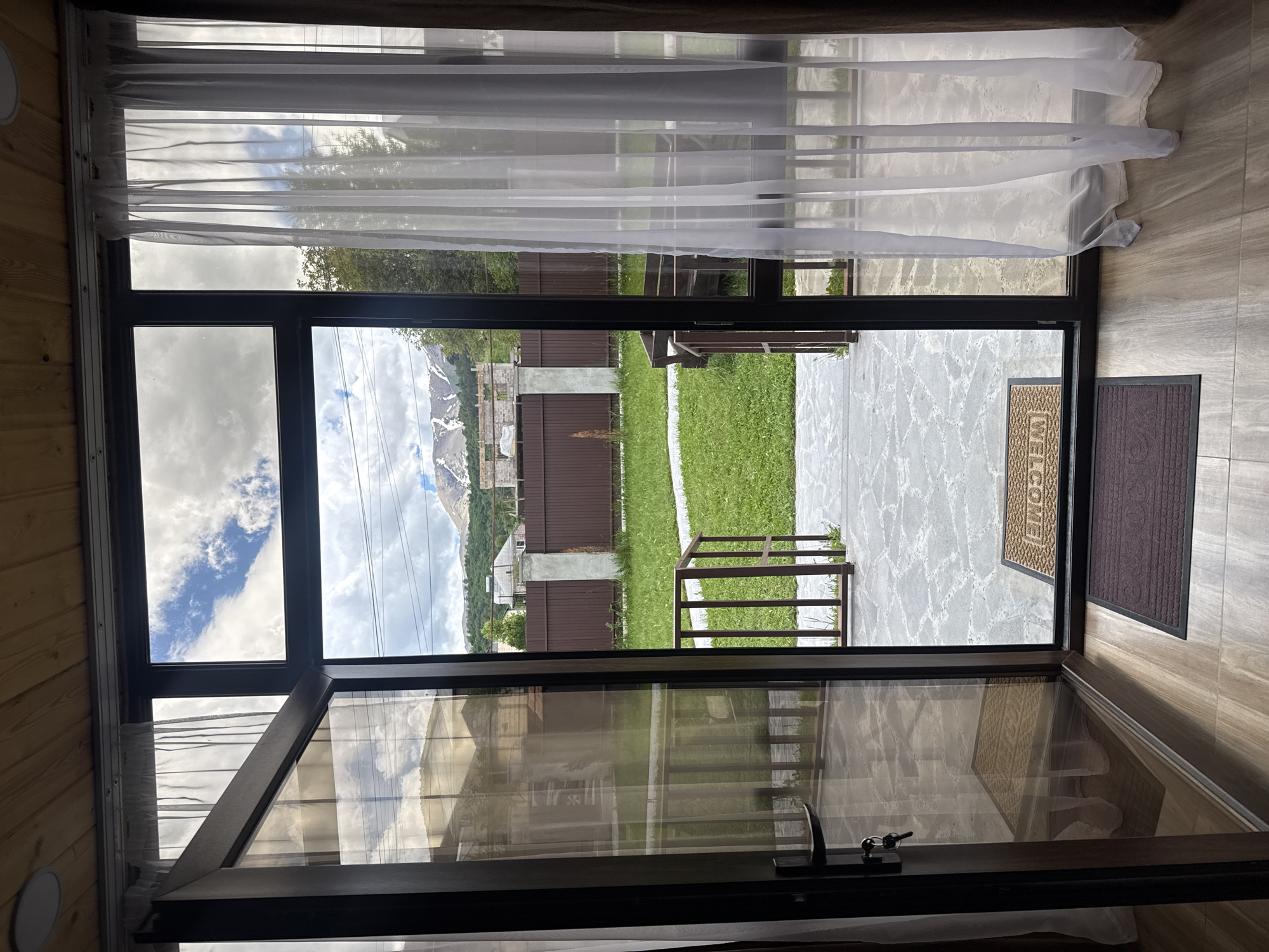 The image depicts a view from inside a building, through a large, open, multi-paneled window or doorway. The interior features wooden walls and flooring. A sheer, white curtain hangs partially covering the upper portion of the window.
Outside, the view encompasses a yard with grass, a concrete pathway, and a small brown metal railing. In the background, there are fences, houses, and a mountain range with visible snow cover on the peaks. The sky is partially cloudy with patches of blue visible. Two mats are visible near the open doorway, one with the word "WELCOME" on it. The scene suggests a location with residential properties and a natural mountain landscape. The mountain range and architecture suggest the location may be near or within the city of Stepanavan, Armenia.