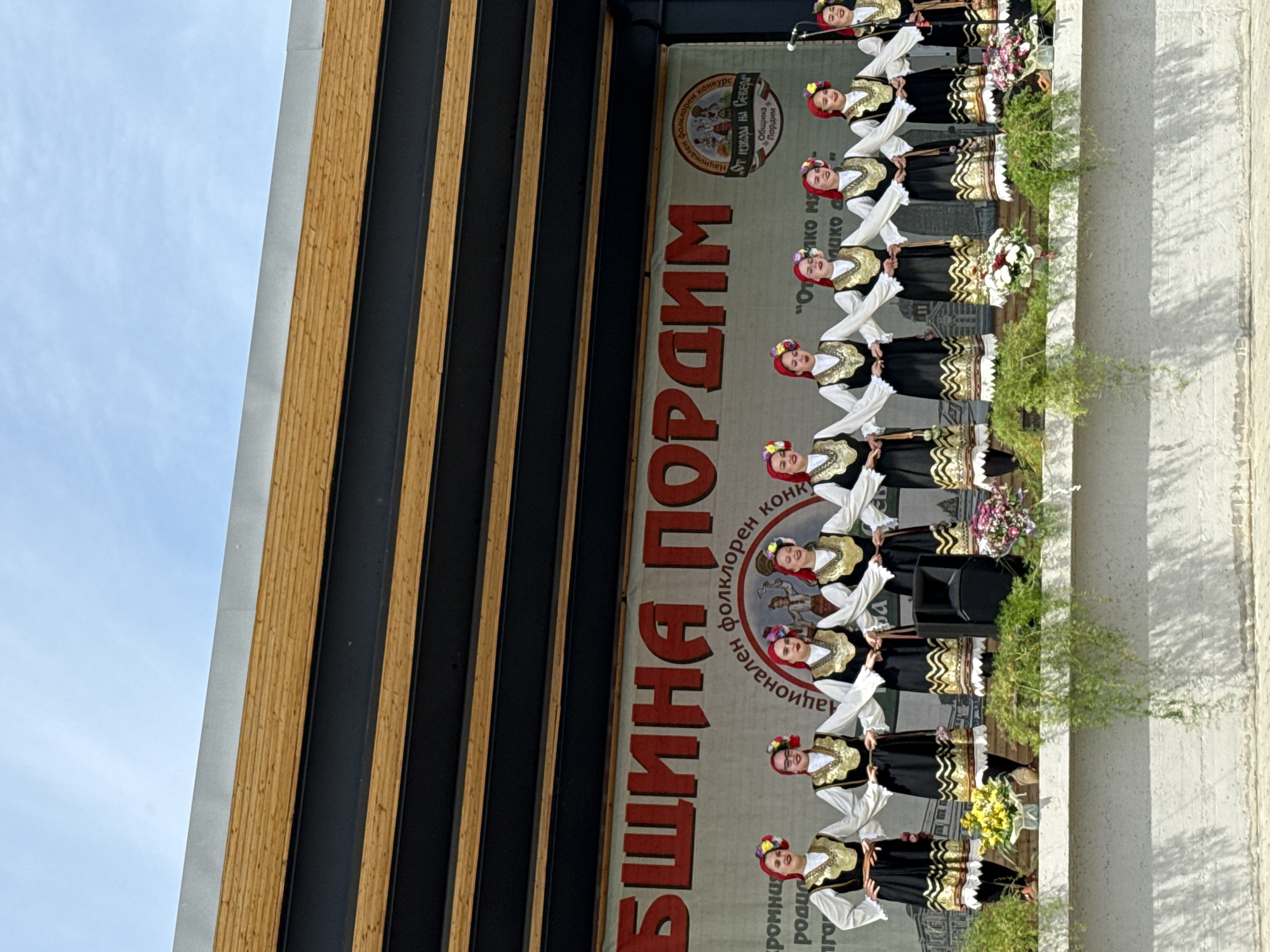 A group of young women in traditional Bulgarian folk costumes are performing on an outdoor stage in Pordim, Bulgaria. The women are arranged in a line, facing forward, and are holding hands. They are wearing white blouses with black vests, embroidered with gold trim, and red headscarves adorned with flowers. The stage backdrop features a large red banner with text in Bulgarian, likely indicating the name of the event or location. To the right of the banner, a circular logo with text and an image is visible. The stage is decorated with floral arrangements. In the foreground, green foliage hangs over the edge of the stage. The sky above is bright and clear, suggesting a sunny day. The scene appears to be a cultural festival or performance, possibly celebrating local traditions and heritage in the municipality of Pordim.