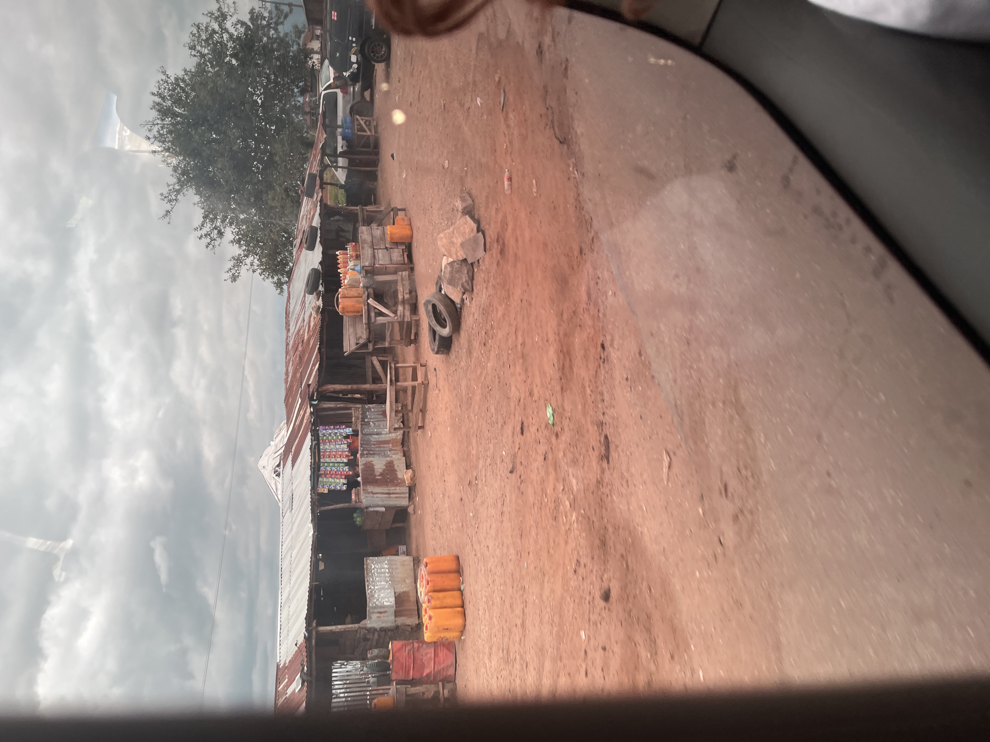 The image depicts an outdoor marketplace in Karshi, Nigeria, characterized by a row of makeshift stalls with corrugated metal roofs. The ground is composed of reddish-brown soil, typical of the region, and scattered with debris including plastic bottles and discarded tires. Several orange plastic containers, likely holding fuel or water, are prominently displayed. Behind the stalls, a large, leafy tree provides some shade, and various vehicles, including a black SUV and a silver van, are parked. The sky is overcast with a dramatic cloud formation. A portion of a person's arm and hair are visible in the lower right corner, suggesting the photograph was taken from inside a vehicle.