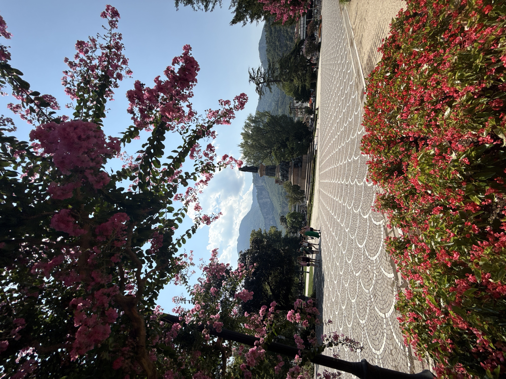 A wide shot captures a sunny day in Trento, Italy, focusing on the Piazza Duomo. In the foreground, vibrant red flowers bloom abundantly. A stone-paved plaza with a concentric circular pattern stretches into the midground, where a monument with a statue stands prominently. Several people, dressed in casual summer attire, are seen walking and conversing on the plaza. Behind the plaza, lush green trees and distant mountains form the backdrop, under a clear blue sky dotted with scattered white clouds.