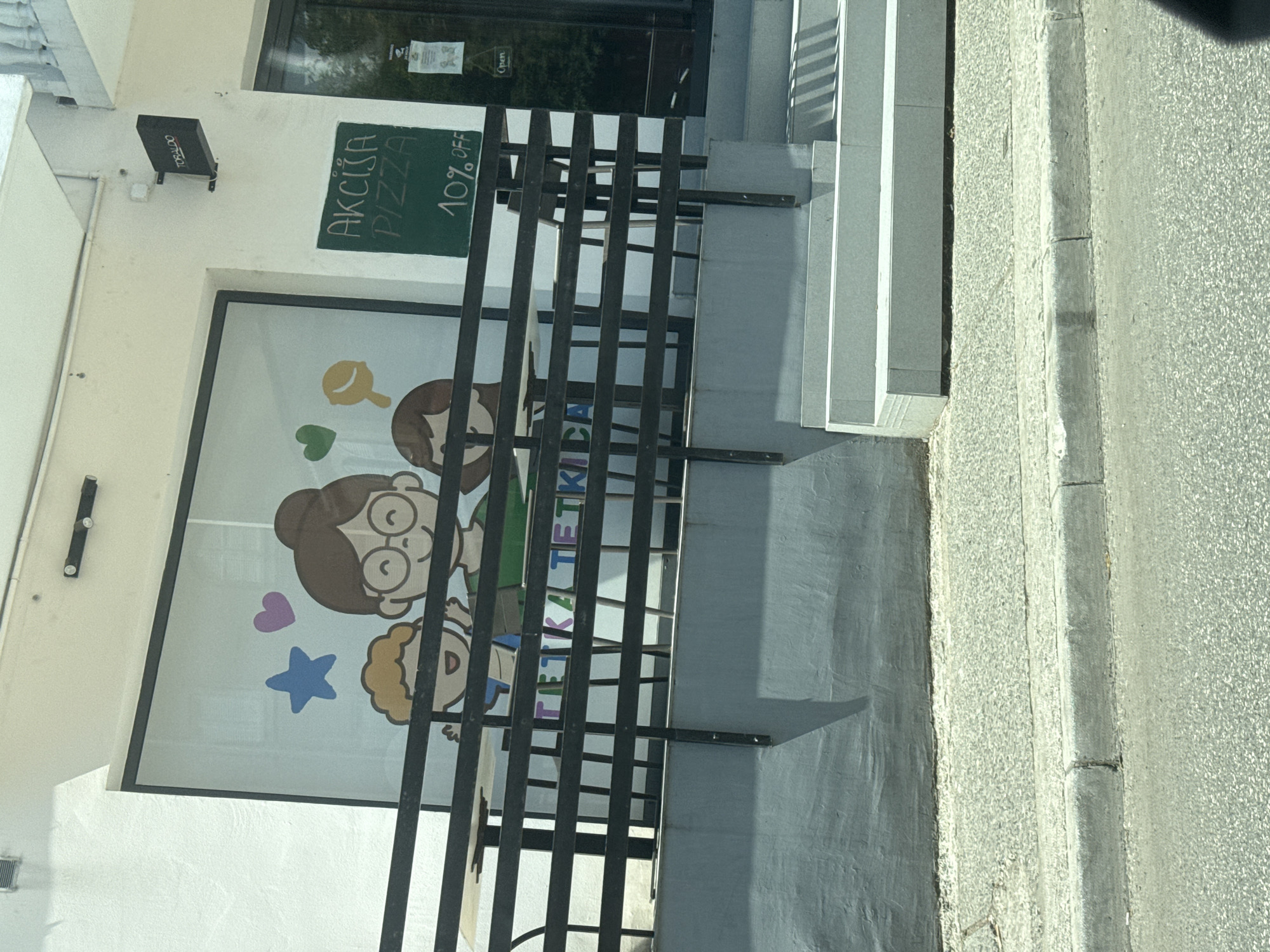 The image shows the storefront of a business, likely a restaurant or cafe, in Budva, Montenegro. The facade is white, with a large window displaying a cartoon illustration of three people. Below the window is a black metal railing, behind which tables and chairs are visible. To the right of the window is a green chalkboard sign advertising "AKCIJA PIZZA 10% OFF". Above the window, a black sign with white lettering reads "TORALDO". The street in front of the establishment is paved, with a sidewalk and a curb. The overall impression is a casual, family-friendly establishment.