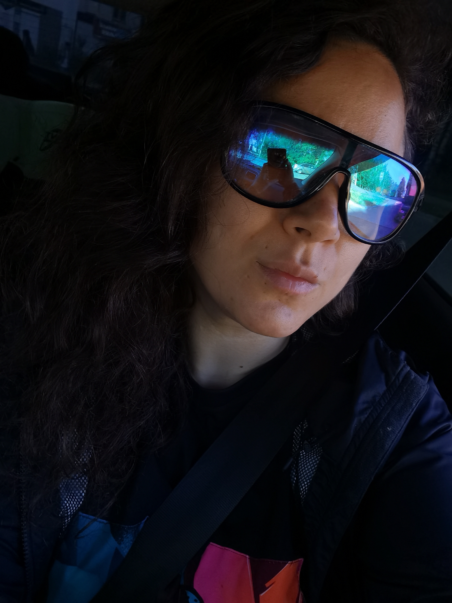 The image shows a close-up, selfie-style shot of a person with long, dark, curly hair wearing large, dark, wraparound sunglasses. The sunglasses reflect a street scene, indicating the person is likely in a vehicle. The reflection shows trees, buildings, and other vehicles. The person is wearing a black t-shirt and a dark vest over it, with a seatbelt visible across their chest. The overall lighting suggests it is daytime, possibly overcast, as the reflections are bright but not harsh. There are no specific landmarks visible that would definitively place the image in Chaikovsky, Russia.