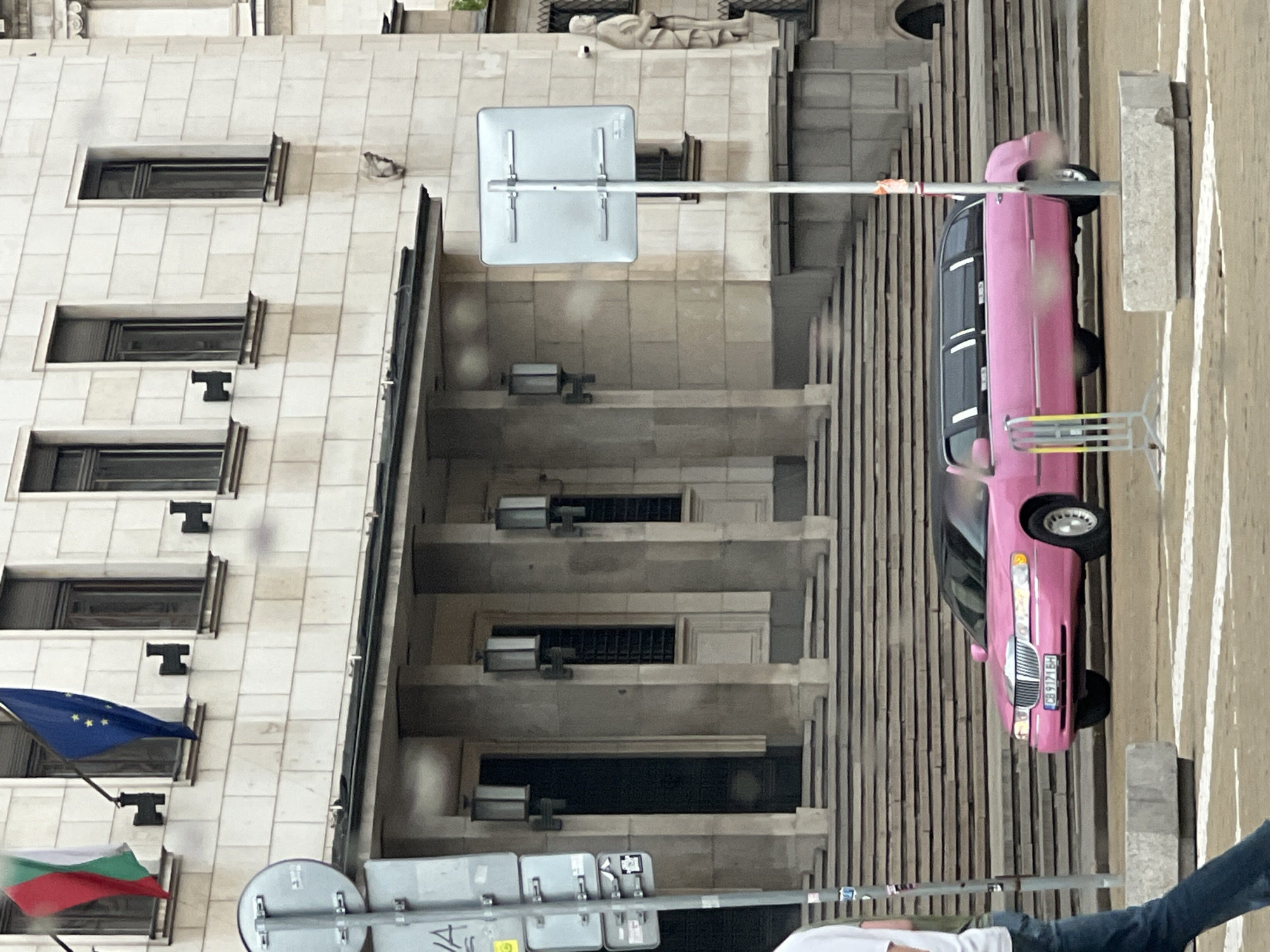The image shows a pink limousine parked on a street in Sofia, Bulgaria, next to a light-colored building with multiple windows and flags, including the Bulgarian and European Union flags. There are also street signs visible. Part of a person wearing a white shirt and jeans is in the foreground. The overall scene appears to be a street view in an urban area.