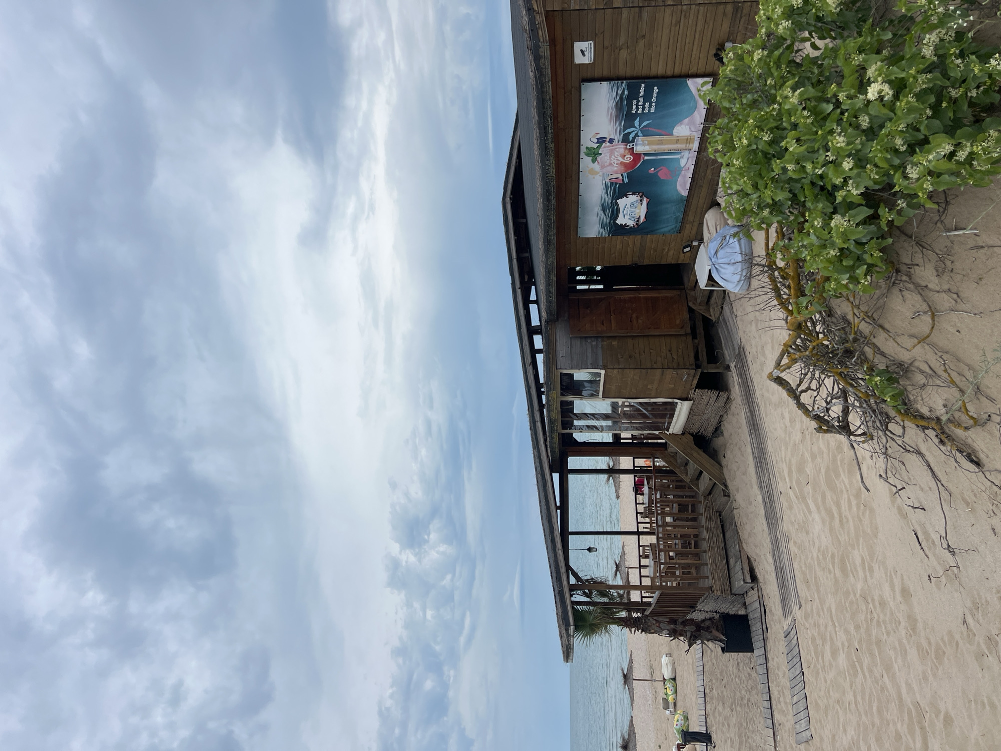 The image shows a wooden beachside restaurant or bar with a covered outdoor seating area. The building has a thatched roof and is situated on a sandy beach. In front of the building, there are tables and chairs. A large banner on the side of the building displays an advertisement for drinks. The sea is visible in the background, with a cloudy sky overhead. There are also beach umbrellas and beanbag chairs on the sand. The overall scene depicts a relaxed, coastal atmosphere. This establishment appears to be located in the city of Primorsko, Bulgaria, as indicated by the presence of a beachside bar and restaurant, common in this resort town.