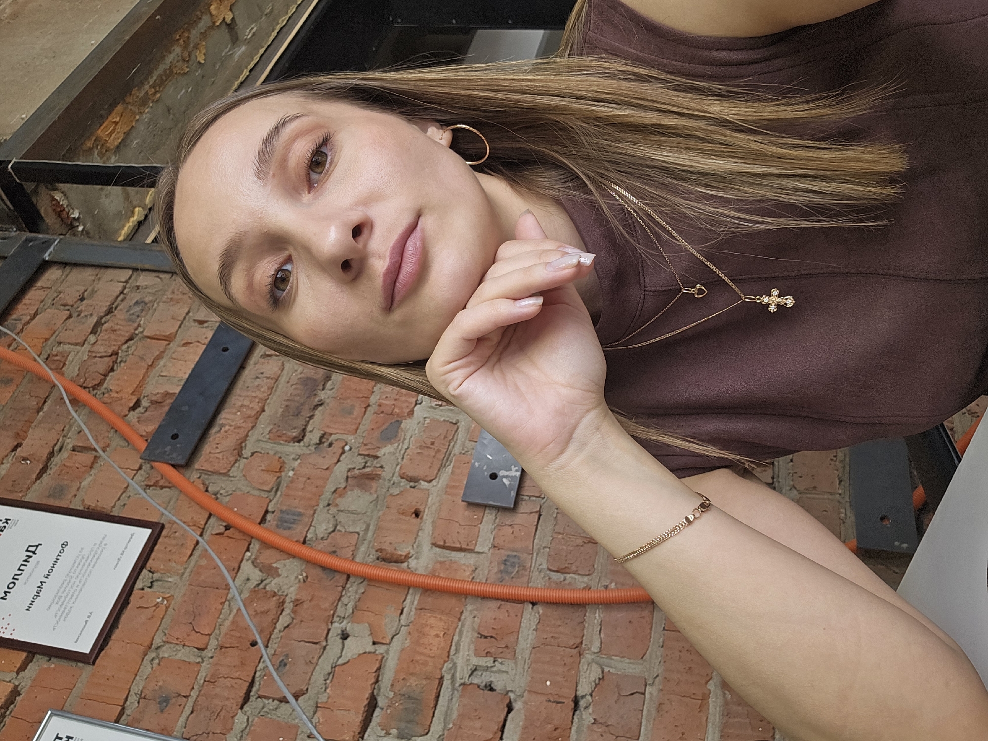 A young woman is posing for a selfie. She has long blonde hair and is wearing a brown sleeveless top, gold hoop earrings, and a gold necklace with a cross pendant. She is resting her chin on her hand, and she is wearing a delicate gold bracelet on her wrist.
The background is a brick wall with an orange pipe running along it. There is also a framed certificate on the wall written in a language other than English. Overhead is an exposed ceiling with metal supports.
The location is Chaikovsky, Russia, based on the document on the wall, but specific building or landmark cannot be determined from the image.