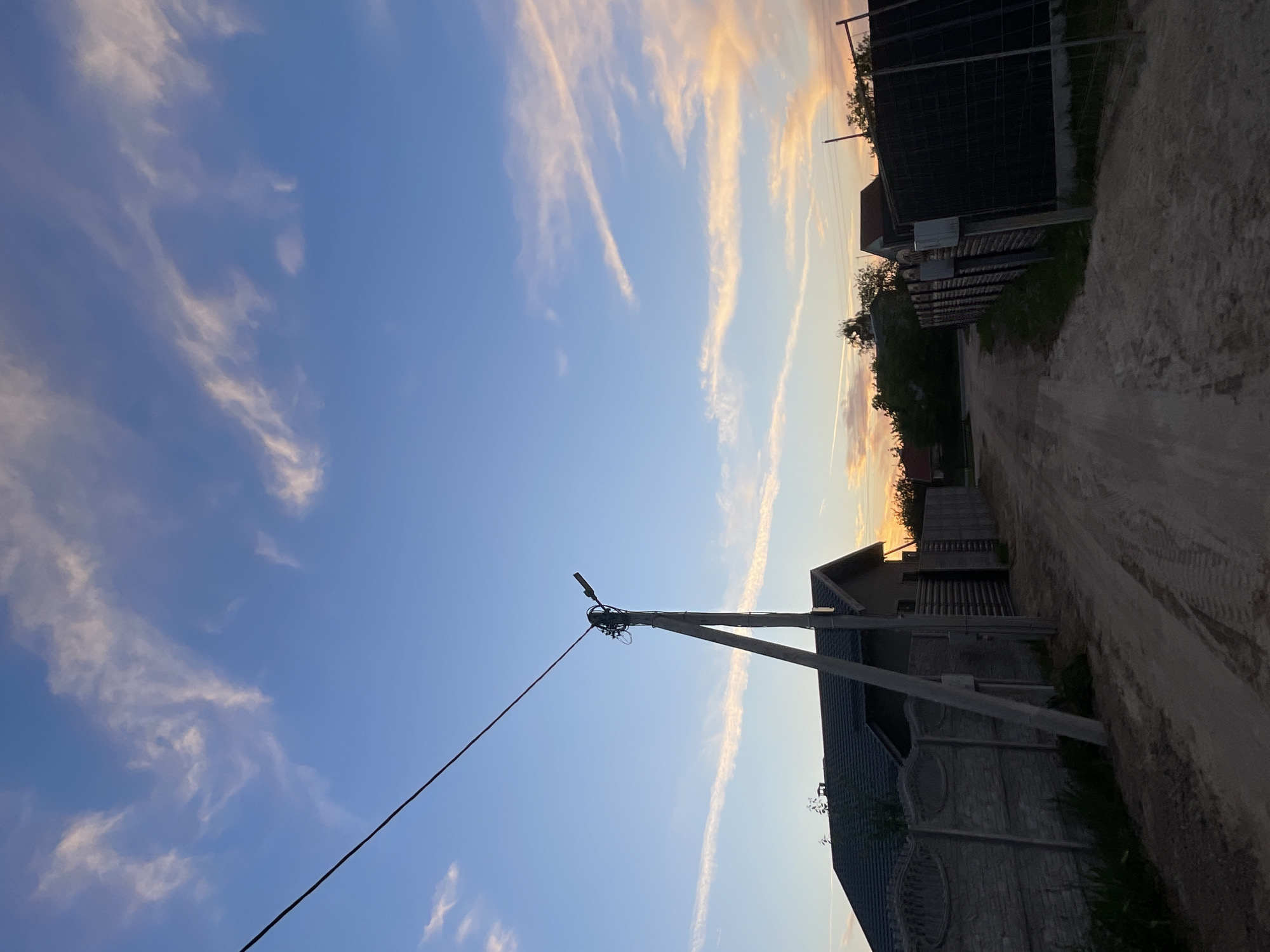 The image depicts a dirt road in a rural area, likely a village or settlement within the Mukhavetsky Selsoviet, Belarus. The sky is a clear blue with streaks of wispy, light pink clouds, suggesting either sunrise or sunset. A prominent concrete utility pole with electrical wires and a street lamp stands on the left side of the road, leaning slightly. On either side of the road are fences, primarily concrete block fences, and some wooden fences. Behind the fences, houses with dark roofs are visible. The road itself is unpaved and shows tire tracks. The overall impression is a quiet, everyday scene in a Belarusian countryside.
