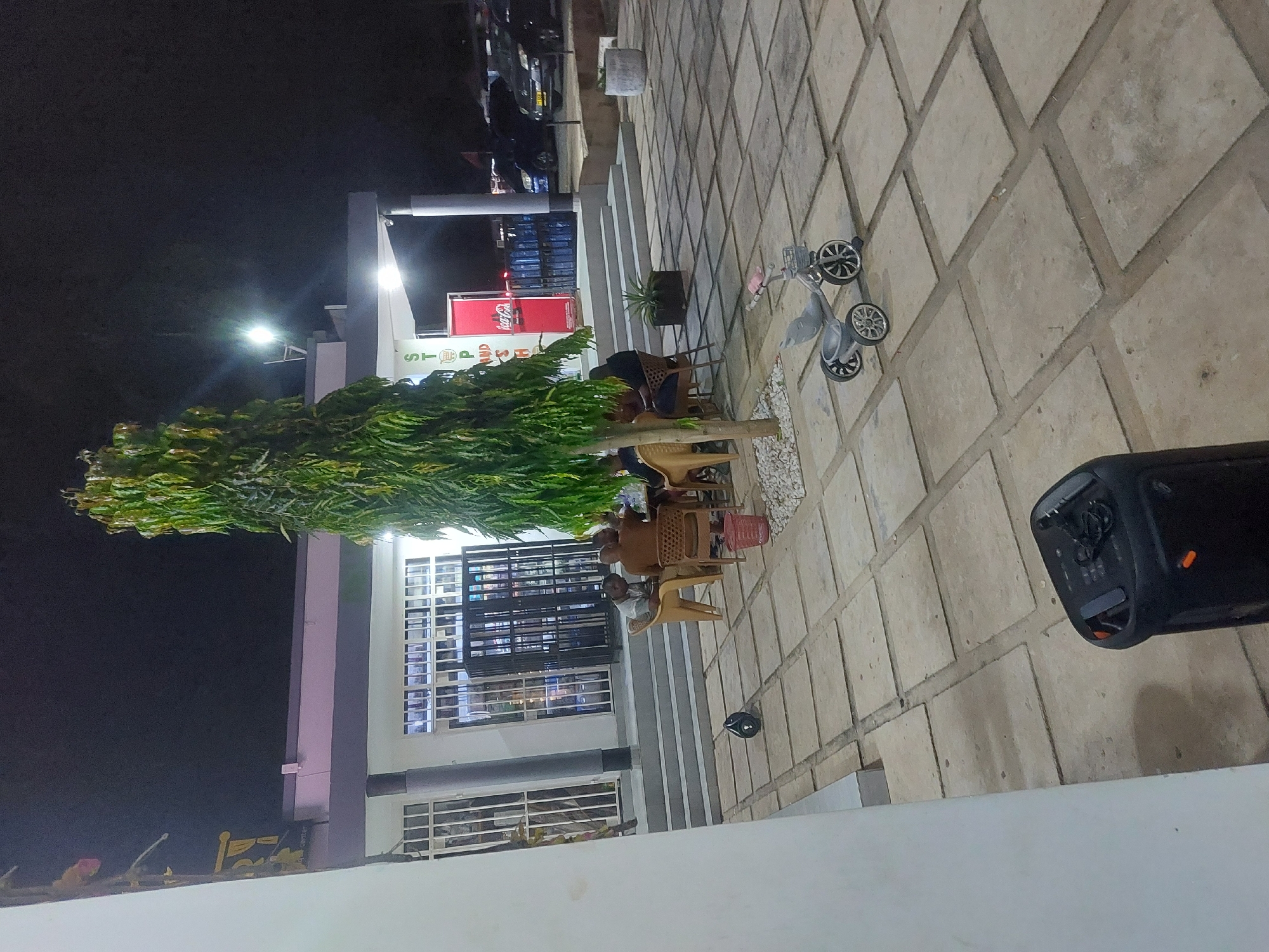 The scene is an outdoor space in Dar es-Salaam, Tanzania, at night. The lighting is artificial, with visible sources. A building with multiple windows and a Coca-Cola sign is present. Several people are seated at tables, likely engaged in conversation. A tall, thin tree partially obscures the view of the building. A gray child's tricycle rests on the paved ground. A black speaker is positioned on the right side of the frame. The ground is covered in square paving stones.