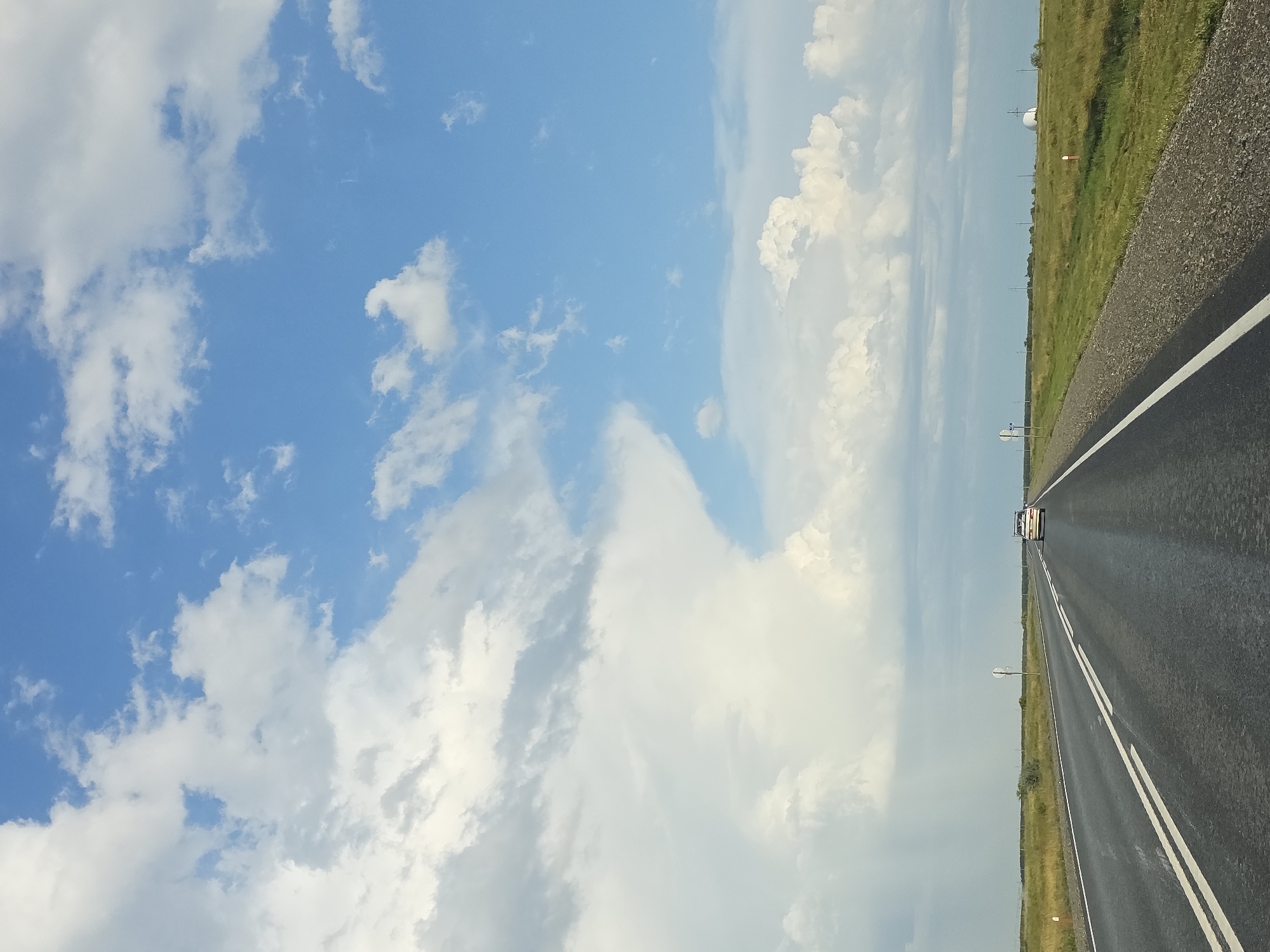 This is a point-of-view shot from a vehicle driving on a two-lane asphalt road in a rural area, likely the Troitsk urban district, Russia. The road has white double solid lines in the center and a white dashed line on the right shoulder. The road surface appears wet, possibly from recent rain. To the right of the road is a grassy field with scattered bushes and a few distant structures, including what appears to be a white domed building. Several utility poles are visible along the horizon. On the left side of the road, there is a grassy verge. The sky is a mix of bright blue and large, fluffy white and gray clouds, with some areas appearing darker and possibly indicating distant rain. A single white car is visible in the distance on the same road, heading away from the viewer. The overall impression is a wide-open, expansive landscape under a dynamic sky.