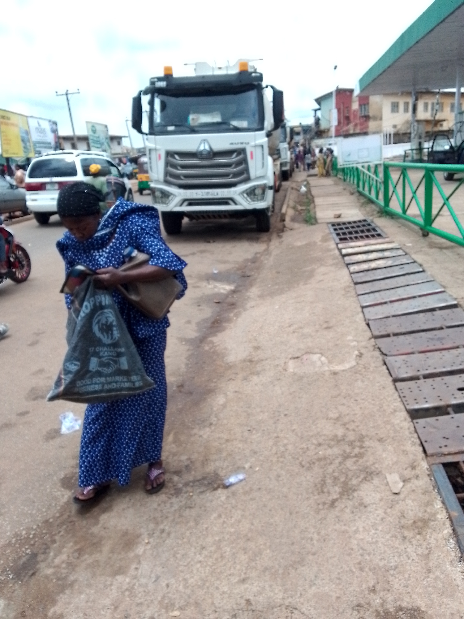 A woman in a blue and white patterned dress and headscarf is holding a black plastic bag with white lettering that says "SHOPPING" and "GOOD FOR MARKETERS BUSINESS AND FAMILIES." She is standing on a dirt road, with a white truck, a white van, and other vehicles in the background. The scene appears to be a busy street in Jos, Nigeria, possibly near a gas station or market. The woman's feet are visible, wearing flip-flops. The overall environment is dusty and worn, with some debris on the ground.