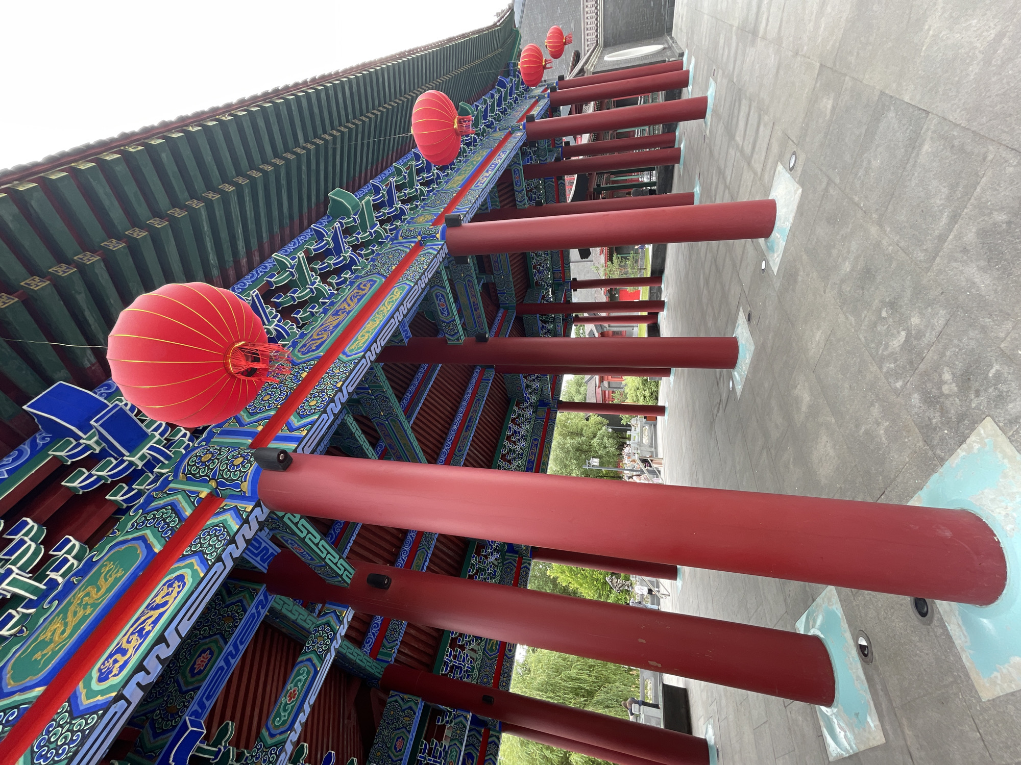 This is a low-angle shot of a traditional Chinese architectural structure, likely a temple or palace, under a cloudy sky. The scene is dominated by the ornate, colorful eaves of the building, featuring intricate patterns in blue, green, gold, and red. Red lanterns hang from the structure, adding a festive touch. Large, bright red pillars support the roof, creating a strong visual line that recedes into the distance. The ground is paved with large, grey stone tiles, and some subtle lighting elements are visible embedded in the paving stones. In the background, glimpses of trees and other structures can be seen, suggesting an outdoor setting. No people are clearly visible, and no text is present in the image. The overall impression is one of grandeur and traditional aesthetics.