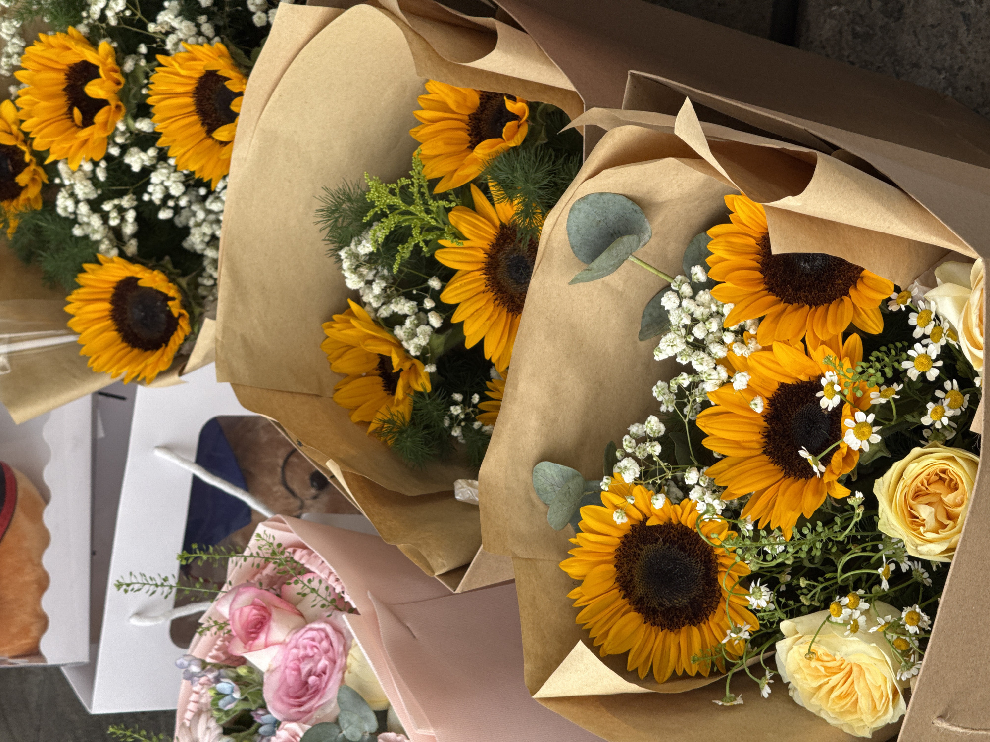 The image shows a collection of floral arrangements, primarily featuring sunflowers, in paper wrapping. The arrangements are presented in brown paper bags, with some also placed in pink and white gift bags. One white gift bag, positioned on the left side of the frame, contains a bouquet of pink roses and other smaller flowers. The background is out of focus but suggests an outdoor setting with a hint of a textured surface. The overall impression is one of a flower shop or market stall. There is no information within the image to suggest it is located in Đà Nẵng, Vietnam.