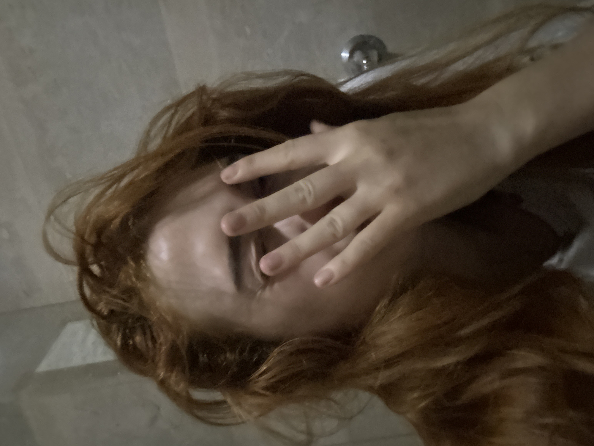 The image shows a person with long, reddish-brown hair lying down on what appears to be a tiled surface. The person's face is partially obscured by their hand. A silver-colored faucet is visible in the background. There is no context to suggest this photo was taken in Budva, Montenegro.