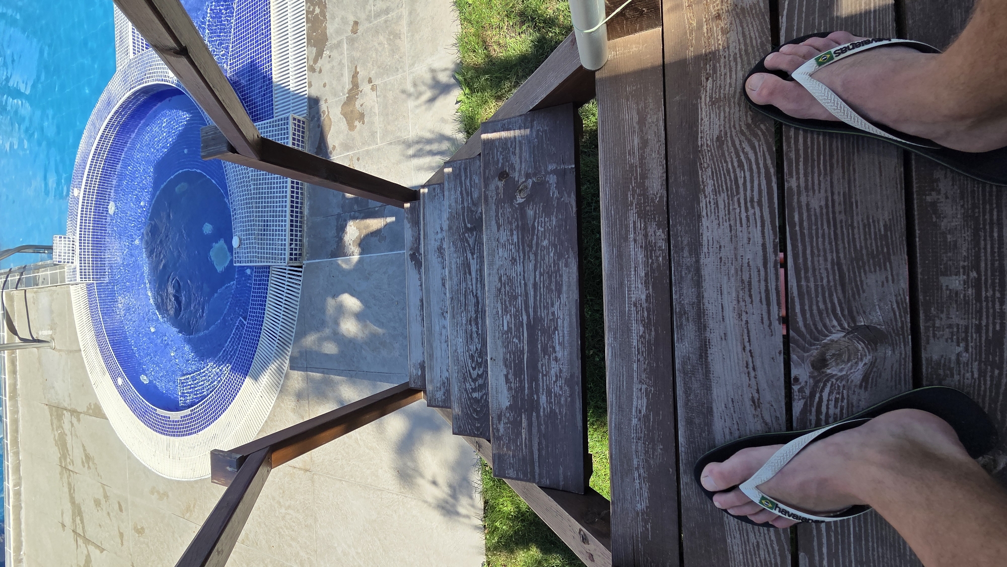 Here is a description of the image:
The image presents a top-down view of a scene that includes a jacuzzi, a set of wooden stairs, and a person's feet in flip-flops. The jacuzzi is circular with blue and white mosaic tiles. Next to the jacuzzi are concrete tiles. Wooden stairs descend from the deck to the jacuzzi area, with a wooden railing on both sides. The individual is standing on the deck near the stairs, wearing black Havaianas flip-flops with white straps. The setting is outdoors. The image appears to be taken during the daytime.