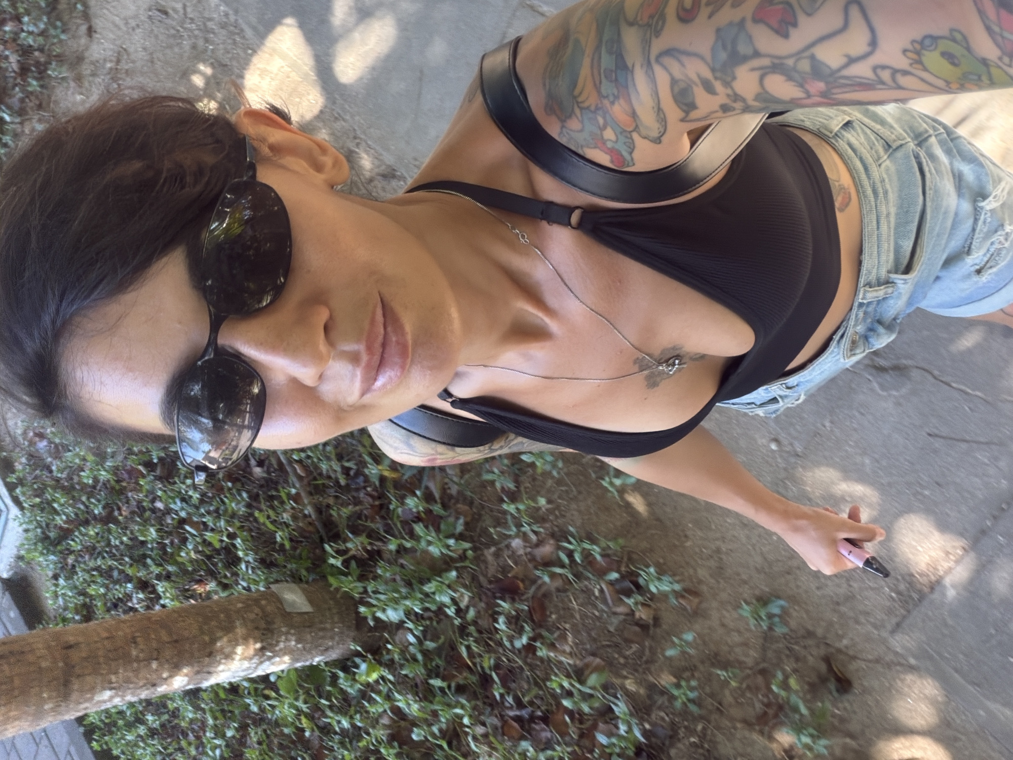 A person wearing sunglasses, a black top, and denim shorts walks on a paved path next to lush green foliage and a tree trunk. The person has tattoos on their arm and is holding a pink object in their right hand. The image appears to be taken in Kuta Selatan, Indonesia.