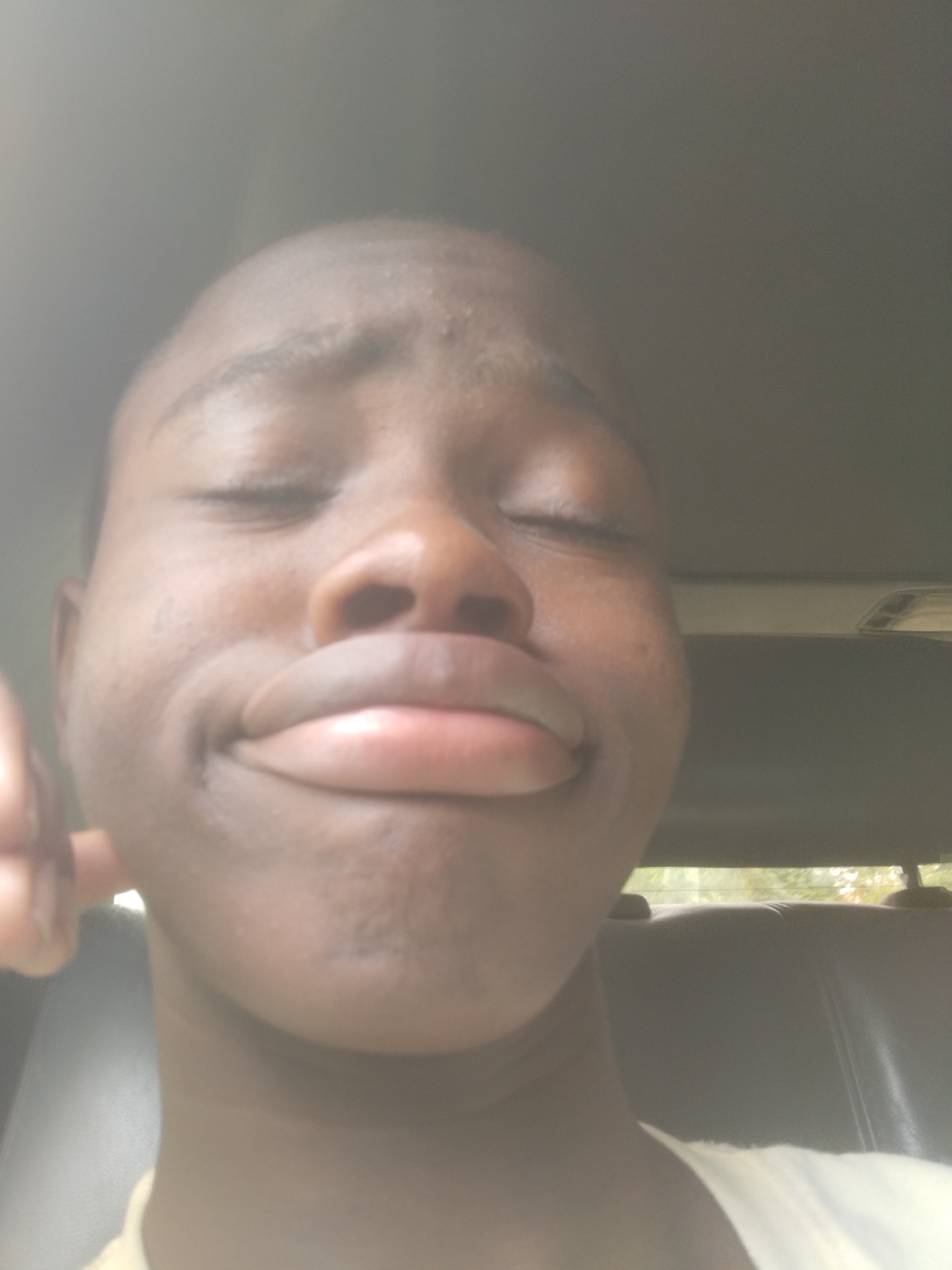 The image shows a close-up selfie of a young, dark-skinned person with their eyes closed and lips puckered. The person appears to be in a car, judging by the interior elements of the vehicle visible in the background, including the seats and a portion of the car's roof. The city and specific location are unknown.