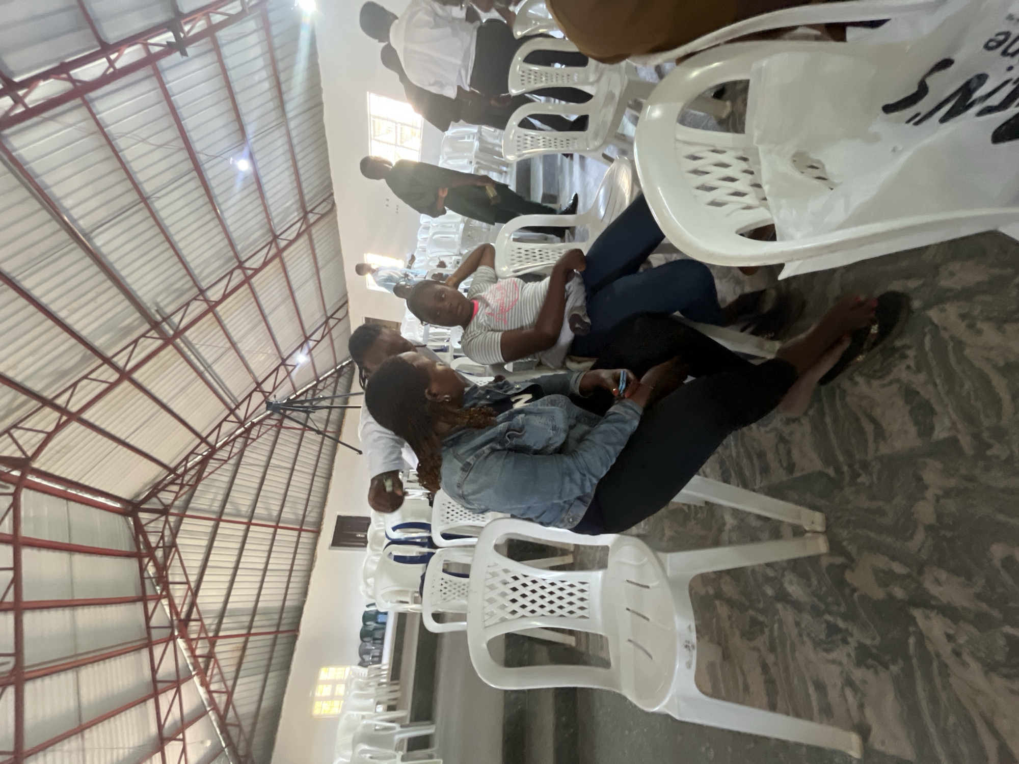 The image shows an indoor scene in what appears to be a large hall in Barakin Akawo, Nigeria. The hall has a high, arched ceiling supported by a metal framework. People are seated on rows of white plastic chairs. Individuals wear a variety of clothing, including casual shirts, denim jackets, and dresses. Some are interacting, while others appear to be waiting or observing. The floor is covered in a marbled-patterned material. Natural light streams in through windows.