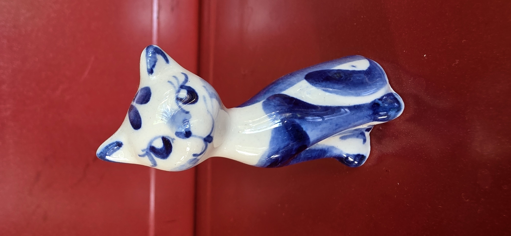 A white ceramic cat figurine with blue floral patterns is placed on a shiny red surface. The figurine is sitting and facing to the left, with its head slightly tilted upwards. The background is a blurred red surface with a slight texture. The image does not contain any information that would allow for localization in the city of Электротехник Снт, Russia.