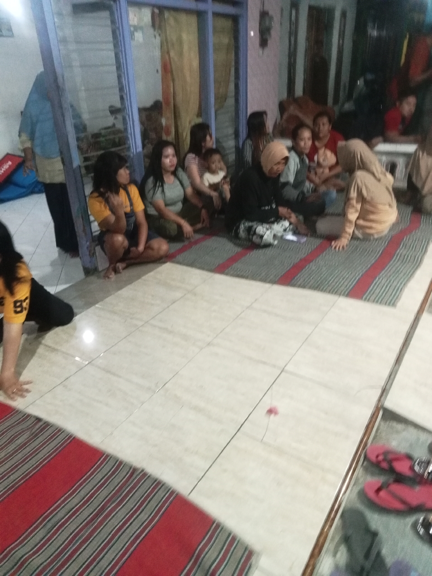 In an indoor setting, likely a living room or communal area, a group of people, primarily women and a few children, are gathered. The flooring is tiled, with a woven mat placed in the center of the gathering. The mat has alternating red and grey stripes. Several individuals are seated on the mat in a relaxed posture, some facing towards a television screen or other focal point not visible in the frame. One woman in the foreground on the left is wearing a yellow shirt with the number "93" on it. Another woman, to her right, is wearing a yellow t-shirt and dark overalls. Other individuals are wearing casual clothing, with some women wearing headscarves. In the background, there's a doorway or window with curtains, and hints of furniture and other household items. The lighting appears to be artificial, casting a slightly dim and warm ambiance. On the right side of the frame, near a reflective surface, a pair of red flip-flops are visible. The overall scene suggests a casual social gathering.