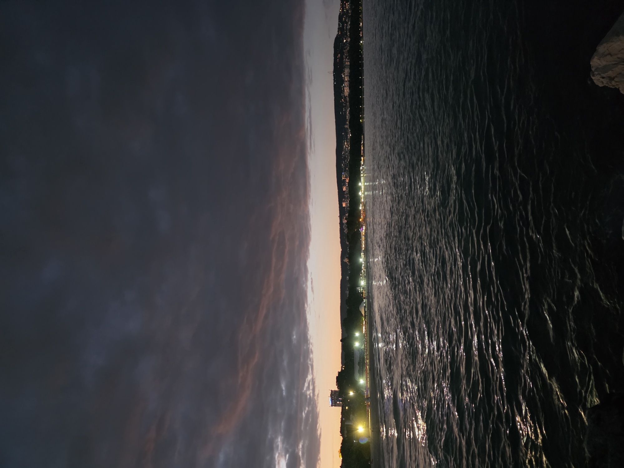 The image depicts a seaside scene at dusk, likely in Varna, Bulgaria. The composition is vertically oriented, with the sky occupying the left half and the sea the right. The sky transitions from dark gray clouds on the far left to lighter hues of orange and pink closer to the horizon, indicating sunset.
Along the horizon, a city skyline is visible, illuminated by numerous lights. These lights twinkle along what appears to be a beachfront promenade or a street bordering the coast. The buildings are silhouetted against the fading light of the sky.
The sea, covering the right side of the image, is dark and appears somewhat choppy, with small waves creating a textured surface. The lights from the city are reflected on the water's surface, creating streaks of brightness. To the far right, a rocky outcrop is visible, adding a natural element to the developed coastline.
The time of day is clearly evening, shortly after sunset, as evidenced by the twilight sky and the city lights being on. The weather appears calm, with no indication of storms or strong winds, although the choppy sea suggests some movement of water. The scene evokes a sense of tranquility and the beauty of a coastal city at nightfall. There is no visible text in the image.