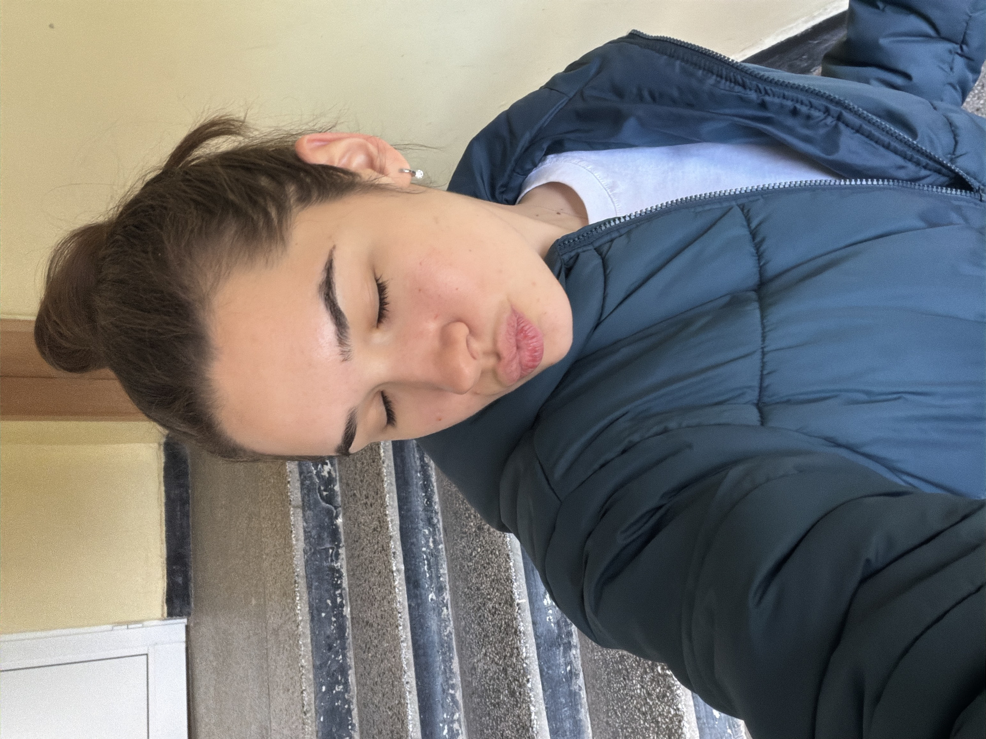 A woman with brown hair pulled back in a bun, wearing a white shirt and a dark blue puffy jacket, is lying down. She is positioned horizontally in the frame, with her head resting on a set of gray, ribbed steps. Her eyes are closed, and her expression is neutral. To the left of the frame is a yellow wall and a portion of a white door. The location is potentially in Sofia, Bulgaria, though there is no clear indicator in the image itself confirming this location.