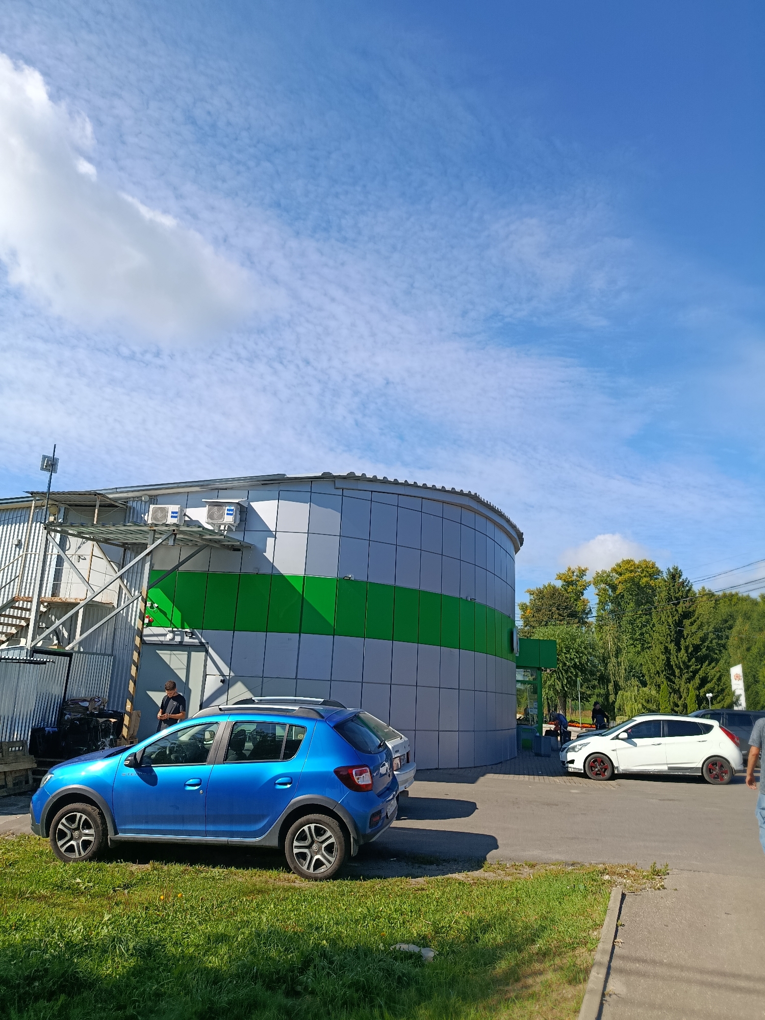 In Vereraya, Russia, on a clear day with scattered clouds, a blue Dacia Sandero is parked on a grassy area next to a paved surface. The car is facing left, and a person is standing near its rear passenger door. Behind the car, a modern, cylindrical building with gray and green panels is visible. An external metal staircase is attached to the left side of the building. Further to the right, a white Hyundai i20 with red rims is parked on the paved surface. A person is seen near the front of this vehicle. In the background, lush green trees and foliage are present. The scene appears to be a commercial or industrial area.