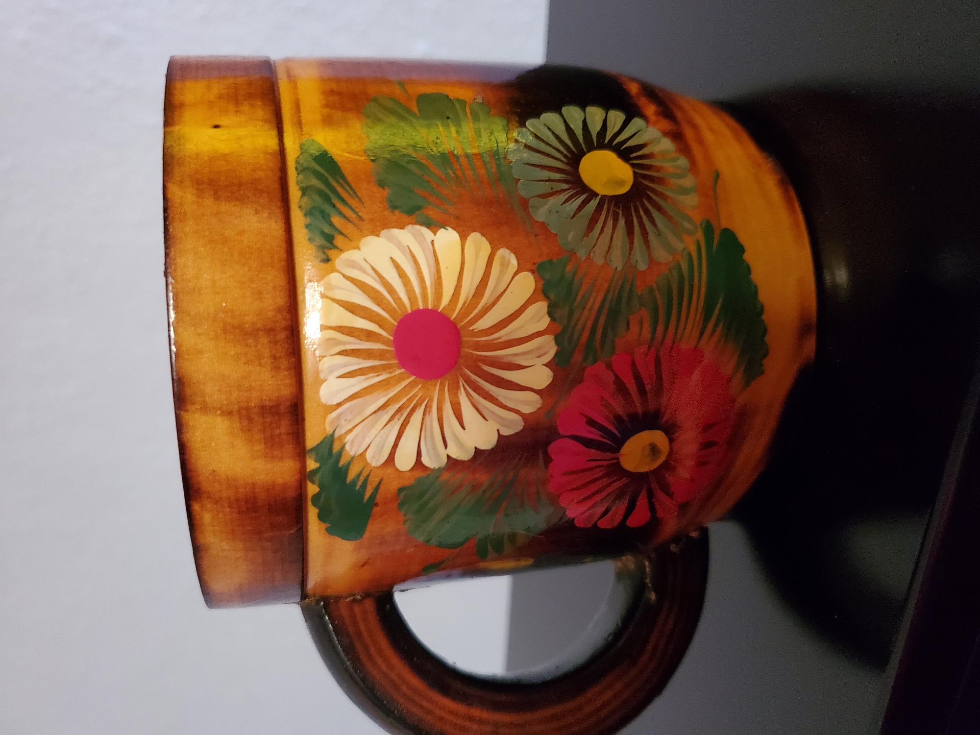 A wooden mug with a handle is decorated with painted flowers and leaves. The mug is a light brown wood with darker brown accents around the rim and handle. The flowers are painted in shades of white, red, and yellow with green leaves. The mug is sitting on a black surface, possibly a table. The background is a plain, light gray wall.