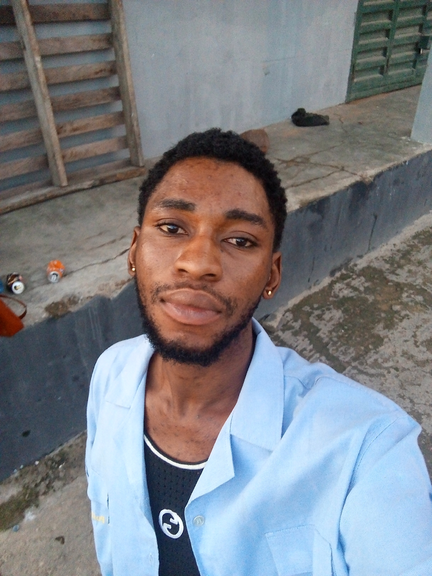 This is a medium shot, selfie-style photograph of a young Black man. He is wearing a light blue button-down shirt over a black tank top with a white circular logo featuring a stylized 'G'. He has short, dark, curly hair and a beard, and is wearing a small gold hoop earring in his left ear. He is looking directly at the camera with a neutral expression.
The background appears to be an outdoor setting in what looks like a residential or urban area. To the left of the man, there is a wooden structure with horizontal planks, possibly part of a fence or stairs. In the middle ground, there are several objects on the ground, including two small colorful cans or bottles, and a dark shoe or slipper. Further back, a gray concrete wall is visible with a closed green shuttered door. The ground is paved with concrete or stone tiles, and appears slightly wet in some areas.
The lighting suggests it is daytime, possibly late afternoon or early morning due to the soft light and shadows. The overall atmosphere is casual and candid. While the location context is Lokoja, Nigeria, there are no specific visual cues within the image that definitively confirm this location. No discernible text is visible in the image.