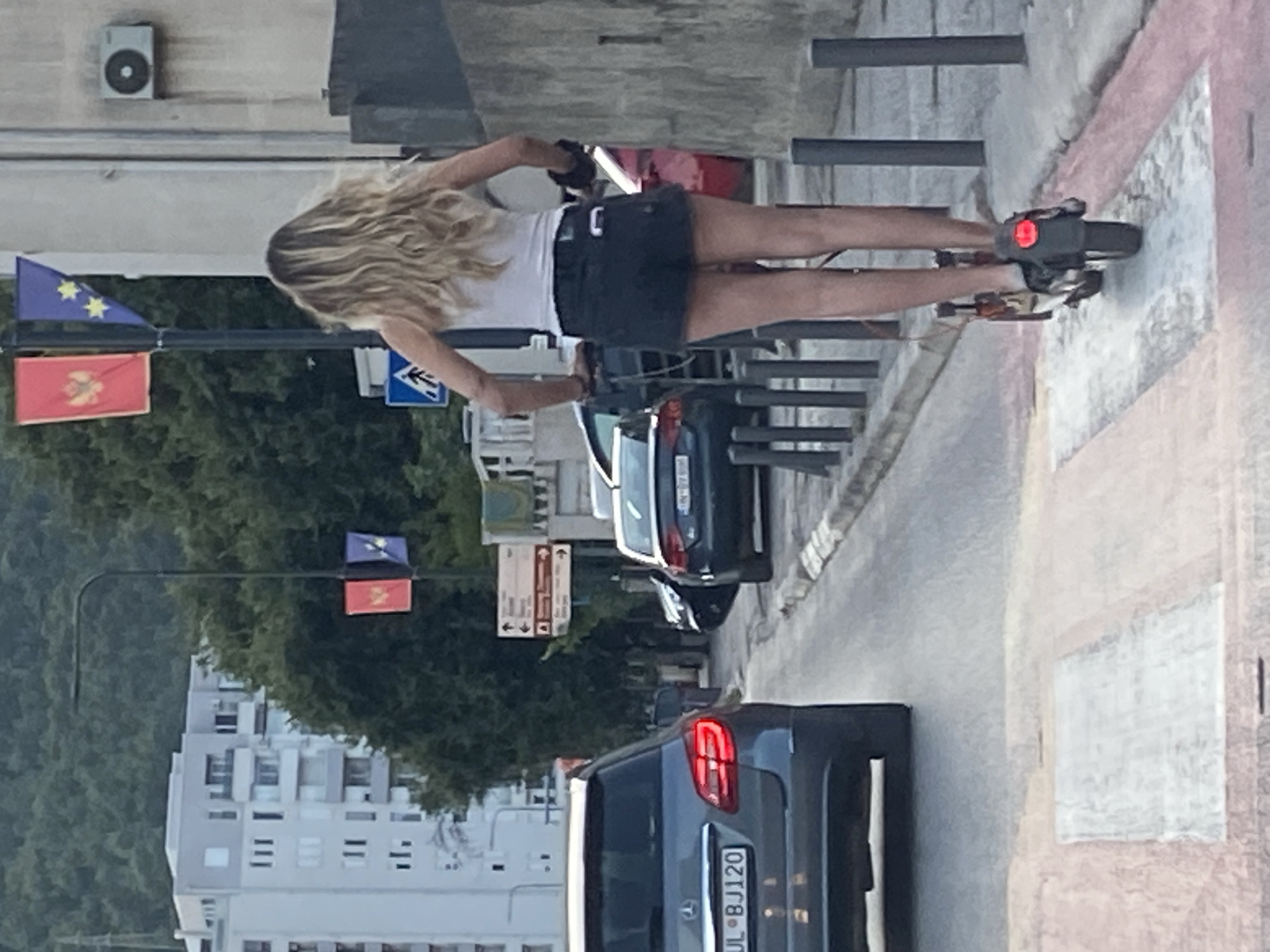 In Budva, Montenegro, on a street with a pedestrian crossing marked by pink and white stripes, a woman with long blonde hair is riding an electric scooter. She is wearing a white tank top and dark denim shorts. Her rear is facing the camera. To her left, a grey Mercedes-Benz SUV is parked. Further down the street, other vehicles and buildings are visible, including a tall apartment building and streetlights adorned with flags. The scene suggests an urban environment with moderate traffic.