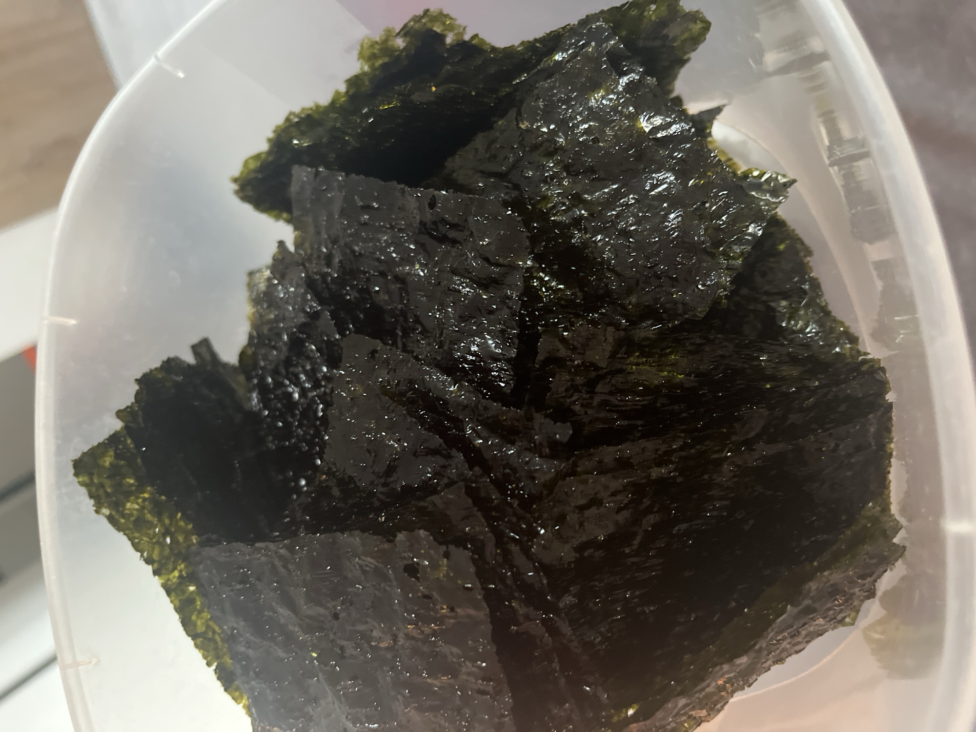 A clear, translucent plastic bowl contains a quantity of dark, irregularly shaped pieces of roasted seaweed, presented in the city of Unknown, Unknown. The seaweed exhibits a prominent oily sheen, reflecting numerous points of light, and a crinkled texture. Its color ranges from deep green to black, with some lighter olive-green hues visible on certain edges and folds. The pieces are loosely stacked within the container, creating varied depths and shadows. The plastic bowl's surface shows minor reflections and its translucency allows for a blurred, light-colored background, likely a tabletop or countertop, to be visible through the material. No individuals, actions, or specific environmental features are discernible beyond the immediate contents and container.