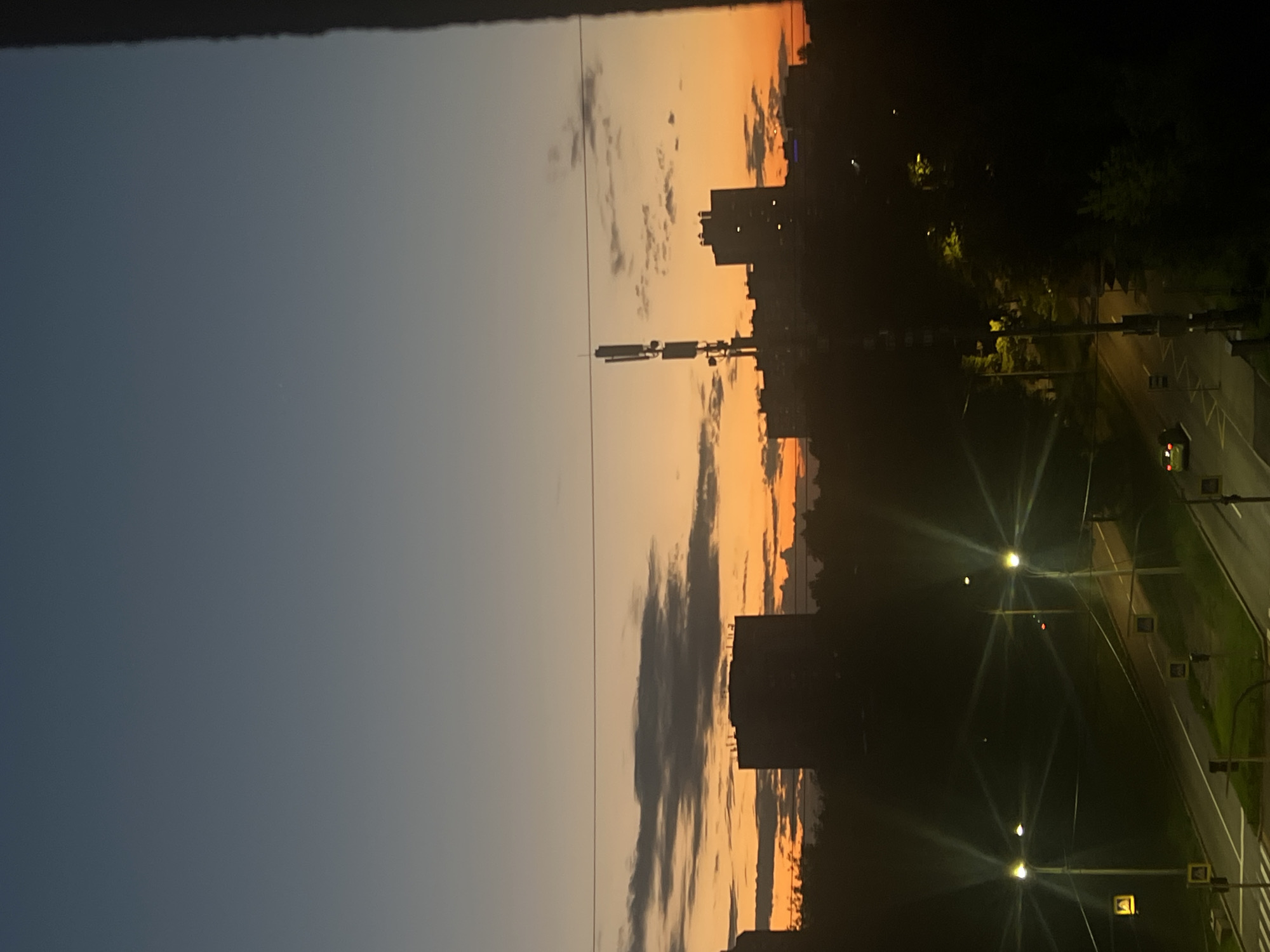 This is a photograph of a city skyline at dusk, likely taken from a high vantage point. The sky transitions from a deep twilight blue at the top to a vibrant orange and yellow near the horizon, indicating the sun has recently set. Wispy clouds are scattered across the sky, catching the last rays of sunlight.
Silhouetted against the colorful sky are several buildings, including apartment blocks and a prominent telecommunications tower with multiple antenna arrays. Trees and other vegetation are visible in the foreground, creating a dark, shadowy layer between the viewer and the illuminated sky.
Below the skyline, streetlights illuminate a road or intersection. Several streetlights are visible, casting distinct starburst patterns of light. Pedestrian crossing signs and road markings are also discernible, suggesting an urban environment. A few vehicles, including a small green car, are present on the road, and their headlights are on.
The scene appears to be a typical urban setting, characterized by residential buildings, infrastructure, and the ambient light of dusk and artificial lighting. Without specific landmarks, it is impossible to confirm the exact location within Saint Petersburg, Russia.
