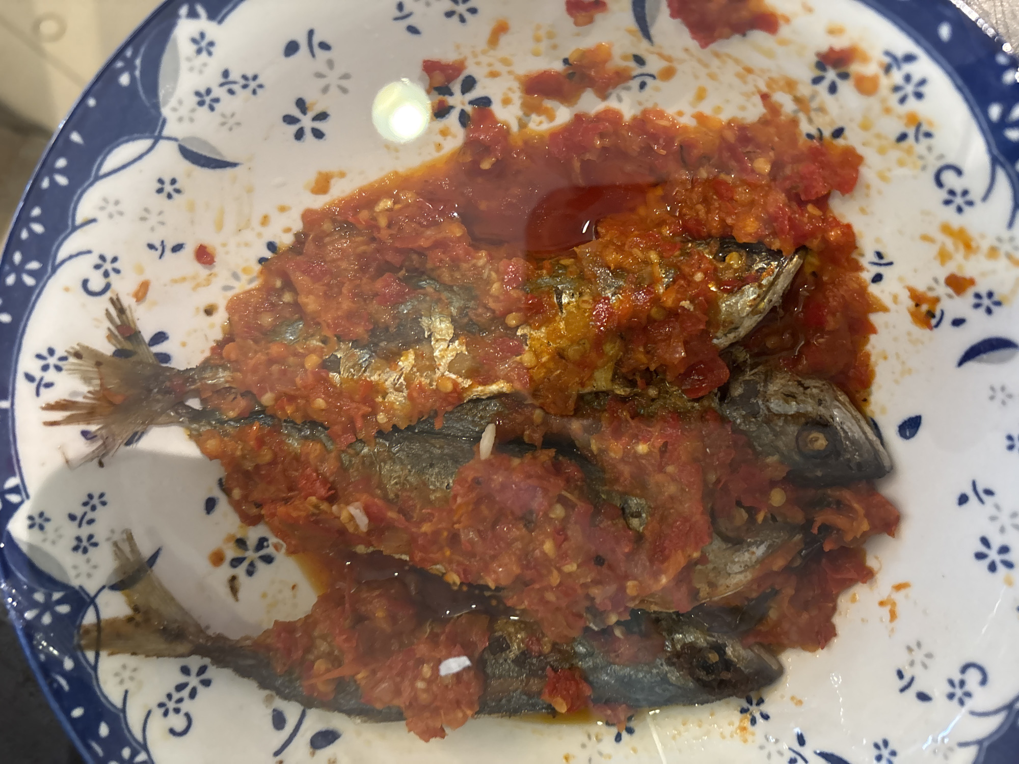 A close-up overhead shot shows three fried fish in a white bowl with blue floral trim. The fish are covered in a red chili sauce. The scene is indoors, likely in a home or restaurant in Pekanbaru, Indonesia.