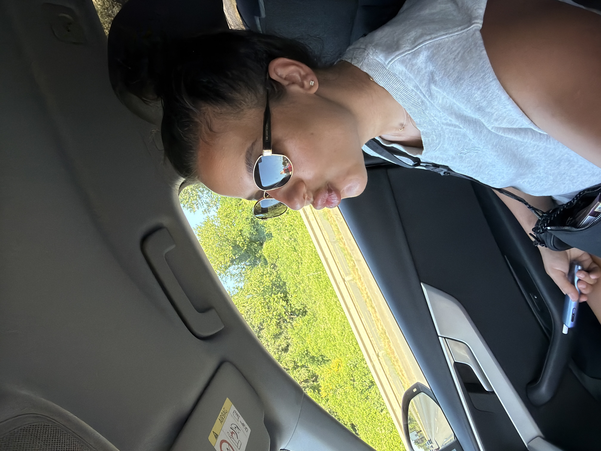 A young woman with dark hair pulled into a bun is seen from the chest up, seated in the passenger seat of a car. She is wearing sunglasses, a light gray t-shirt, and a necklace. Her lips are pursed as if for a kiss, and she is looking to her right. In her right hand, she holds a small, light blue cylindrical object, possibly an e-cigarette. The interior of the car is visible, including the car door panel, a door handle, and the car's headliner with an airbag warning sticker. Through the window, lush green trees and a glimpse of a road with a concrete barrier are visible, suggesting a drive through a scenic or suburban area. The lighting indicates it is daytime, likely sunny. The image does not contain any information to suggest the location is the city of Burgas, Bulgaria.