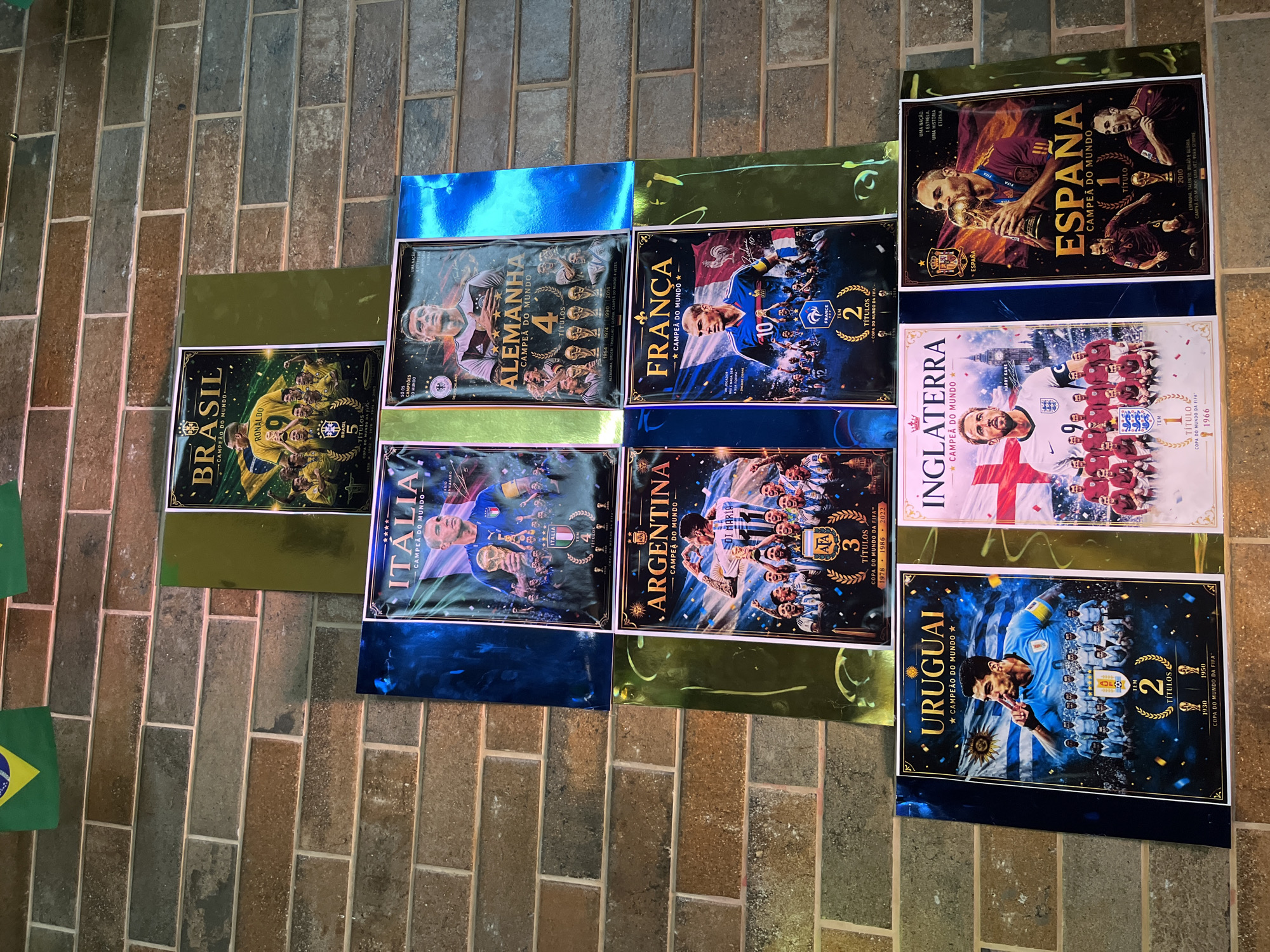 The image shows a collection of posters arranged on a floor with a brick pattern. The posters feature soccer teams that have won the World Cup: Brazil (5 wins), Germany (4 wins), Italy (4 wins), Argentina (3 wins), France (2 wins), Uruguay (2 wins), England (1 win), and Spain (1 win). Each poster has a player or team image and states the nation's name and the number of World Cup titles. The posters are partially bordered by shiny gold and blue paper, suggesting they are possibly being framed or displayed. A small Brazilian flag is visible in the lower left. The location is Unknown, Unknown.