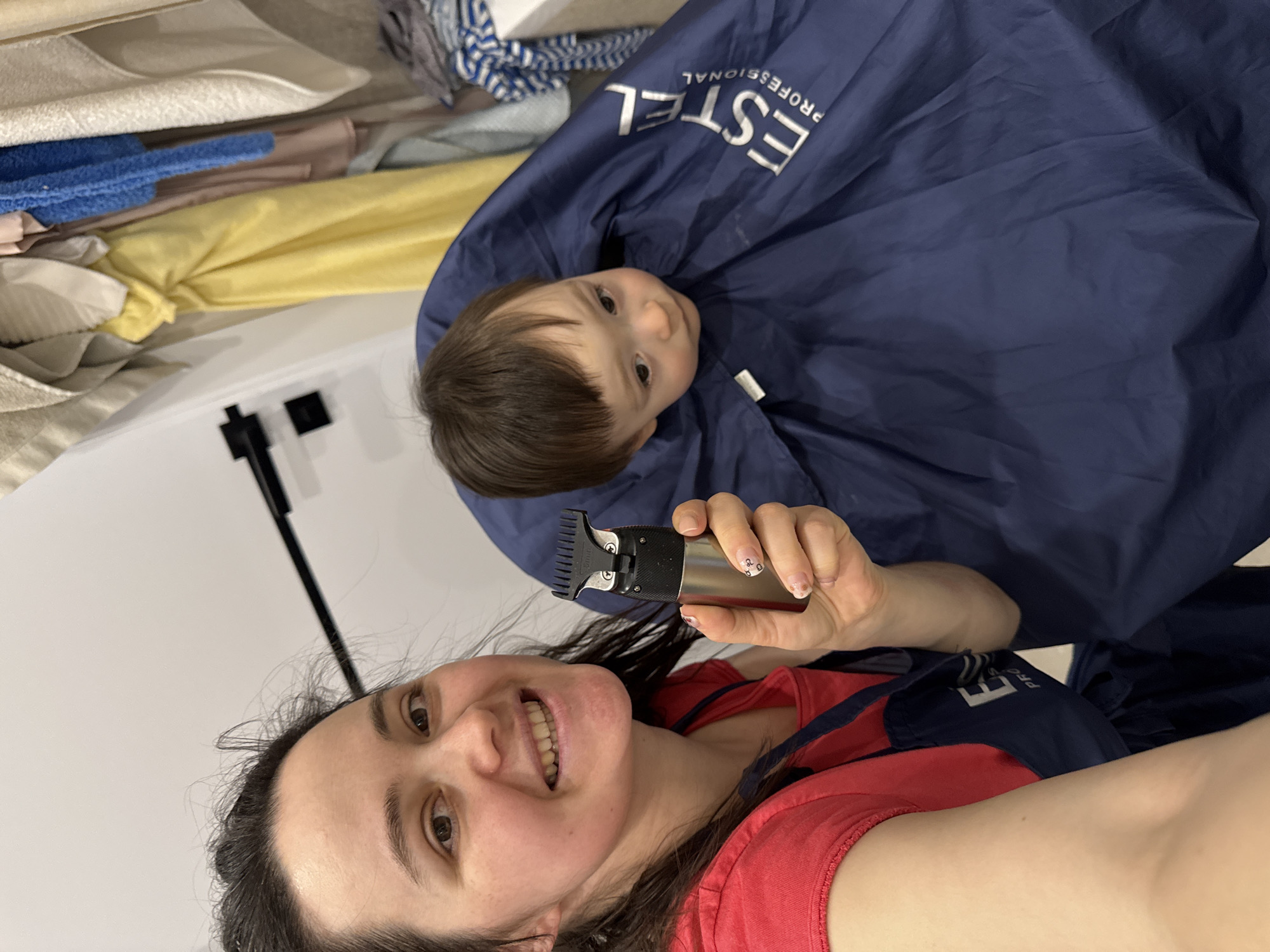 The image is a vertical, selfie-style photograph, rotated 90 degrees counter-clockwise. It features two individuals: a woman and a young child, engaged in what appears to be a home haircut activity.
In the foreground, on the left side of the frame, an adult woman with dark hair and a warm, wide smile looks directly at the camera. She is wearing a red top and a blue barber's smock. In her right hand, she holds a silver and black electric hair clipper, positioned as if ready to use. Her fingernails show light-colored polish with small decorative elements.
To her right and slightly behind, a young child, likely a toddler, looks upwards with a neutral, slightly curious expression. The child is also wearing a blue barber's smock, similar to the woman's. Prominently visible on the child's smock, in white and inverted text, is the brand name "ESTEL PROFESSIONAL." The child's dark hair frames their face.
The background, situated above the subjects in the rotated image, consists of neatly stacked textiles, including towels or blankets in various colors such as white, beige, blue, and yellow. A black pole or stand is also visible against a plain white wall.
The overall scene suggests a joyful and intimate moment during a home hair-cutting session, likely taking place indoors in a domestic environment, possibly in Izhevsk, Russia, given the brand on the smock. The woman's expression conveys happiness and affection.